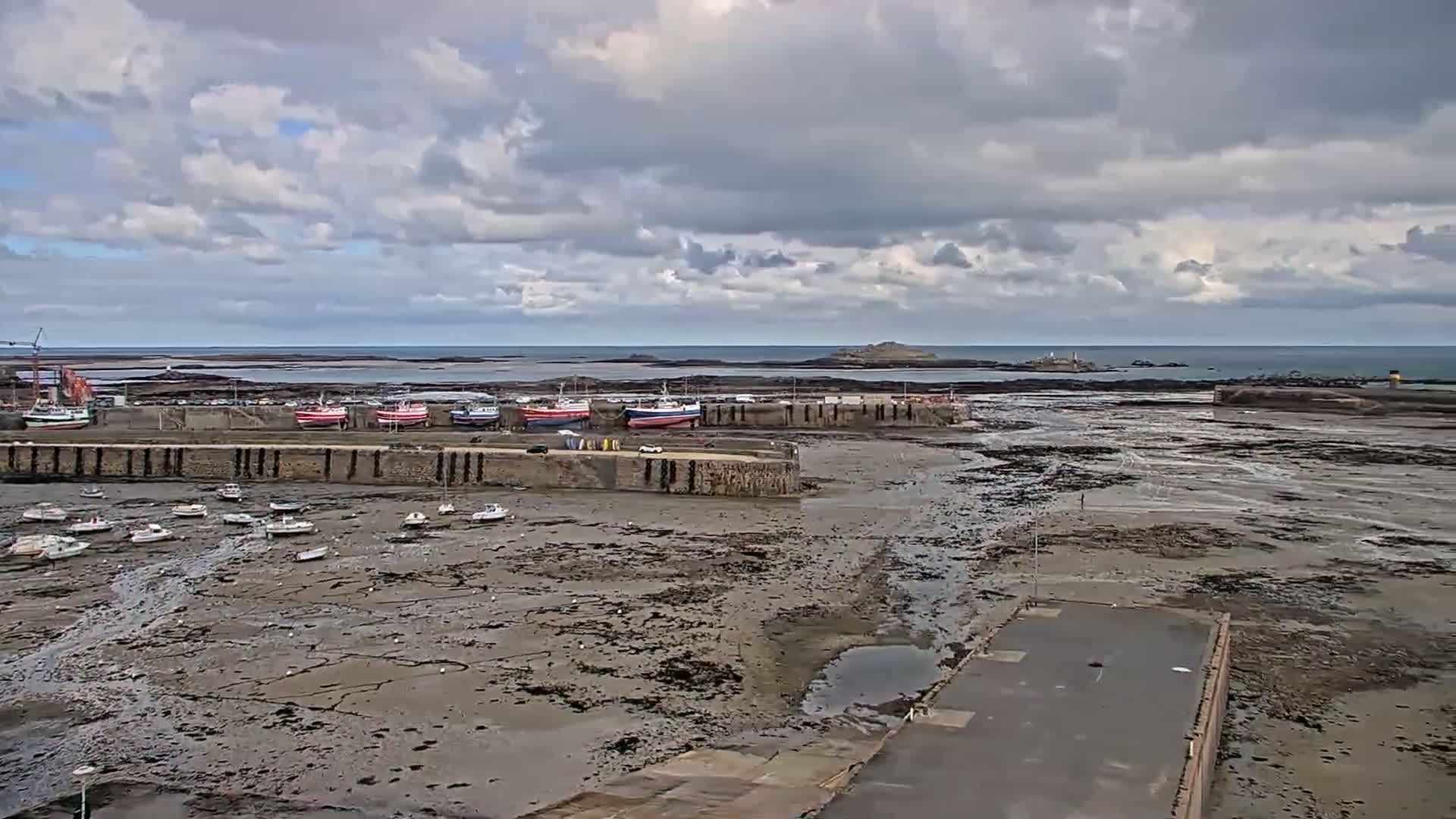 A harbor with numerous small boats rests on exposed mudflats under a cloudy sky, with an island visible in the distance.