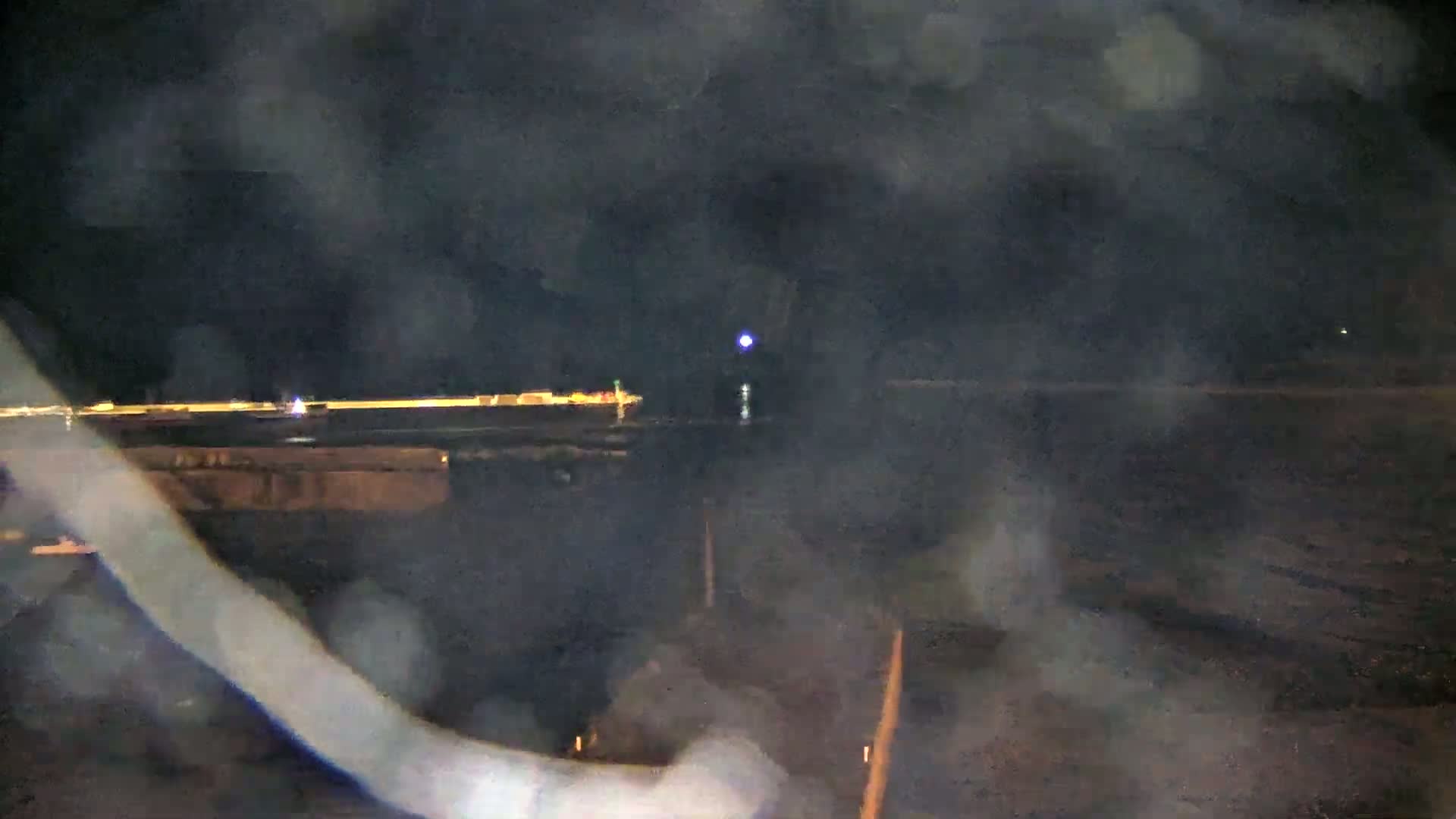 A blurry nighttime view shows a dimly lit, dark area with a line of lights in the distance, possibly a roadway or runway, under a hazy, dark sky.