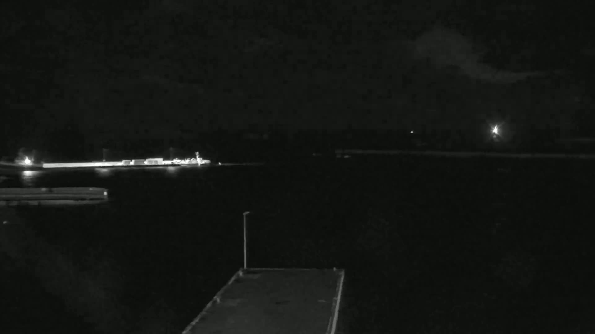 A nighttime, low-light view of a dark body of water with some structures faintly visible on the shore and a single light source in the distance under a cloudy sky.