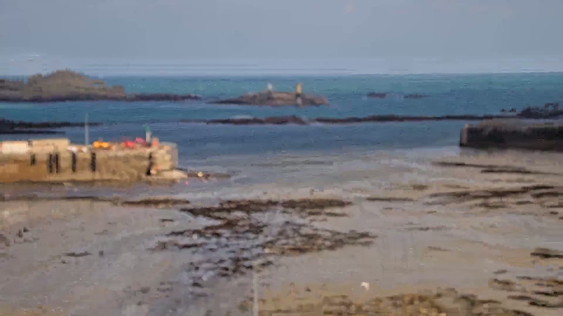 A mostly calm ocean with a low tide revealing a sandy seabed and rocky outcroppings near a stone harbor wall.