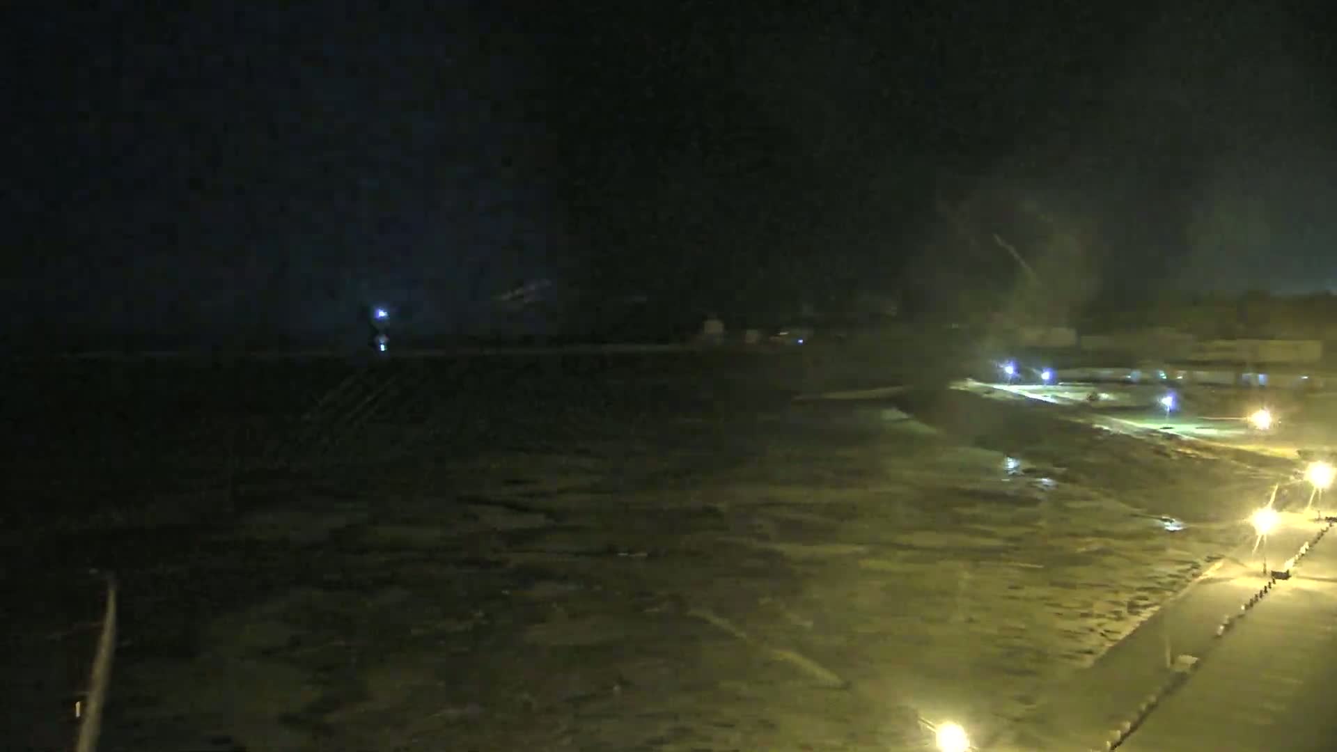 A nighttime, dimly lit coastal scene shows choppy water under a dark sky, with lights illuminating a paved walkway and nearby structures.