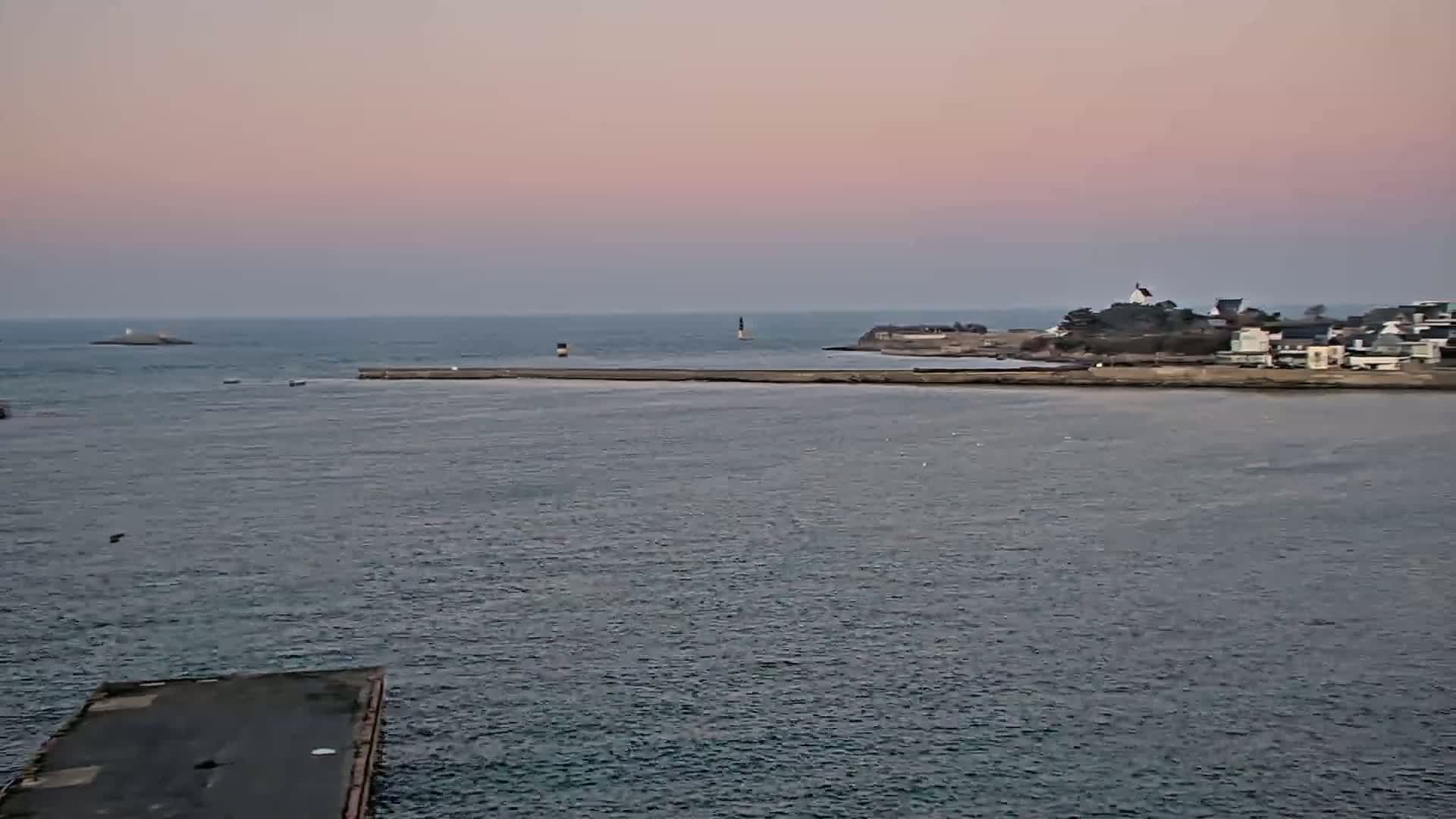 A calm body of water under a pale pink sky shows a small town on the far shore, a breakwater, and a small island in the distance.