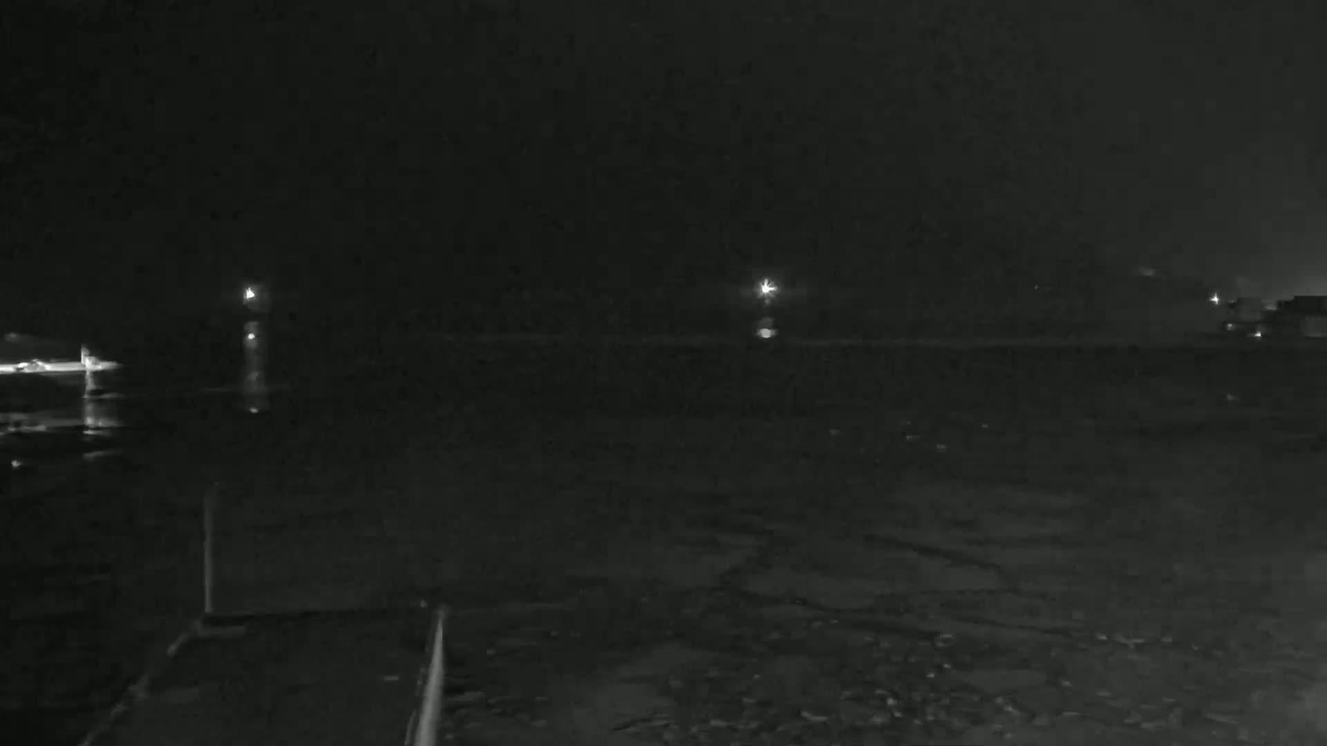 A dark, nighttime scene shows a body of water reflecting a few distant lights under a cloudy sky.
