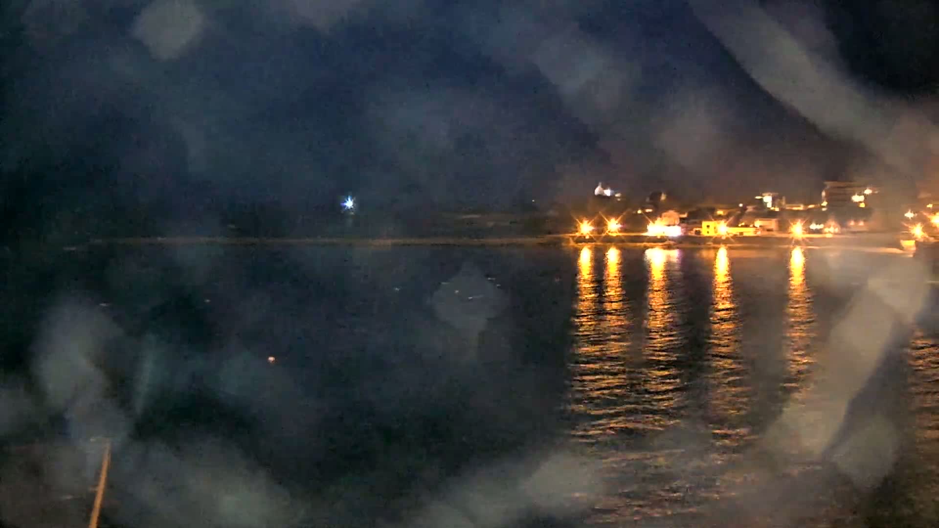 A nighttime view across a body of water shows a shoreline with illuminated buildings and their reflections shimmering on the dark water, viewed through a hazy or misty atmosphere.