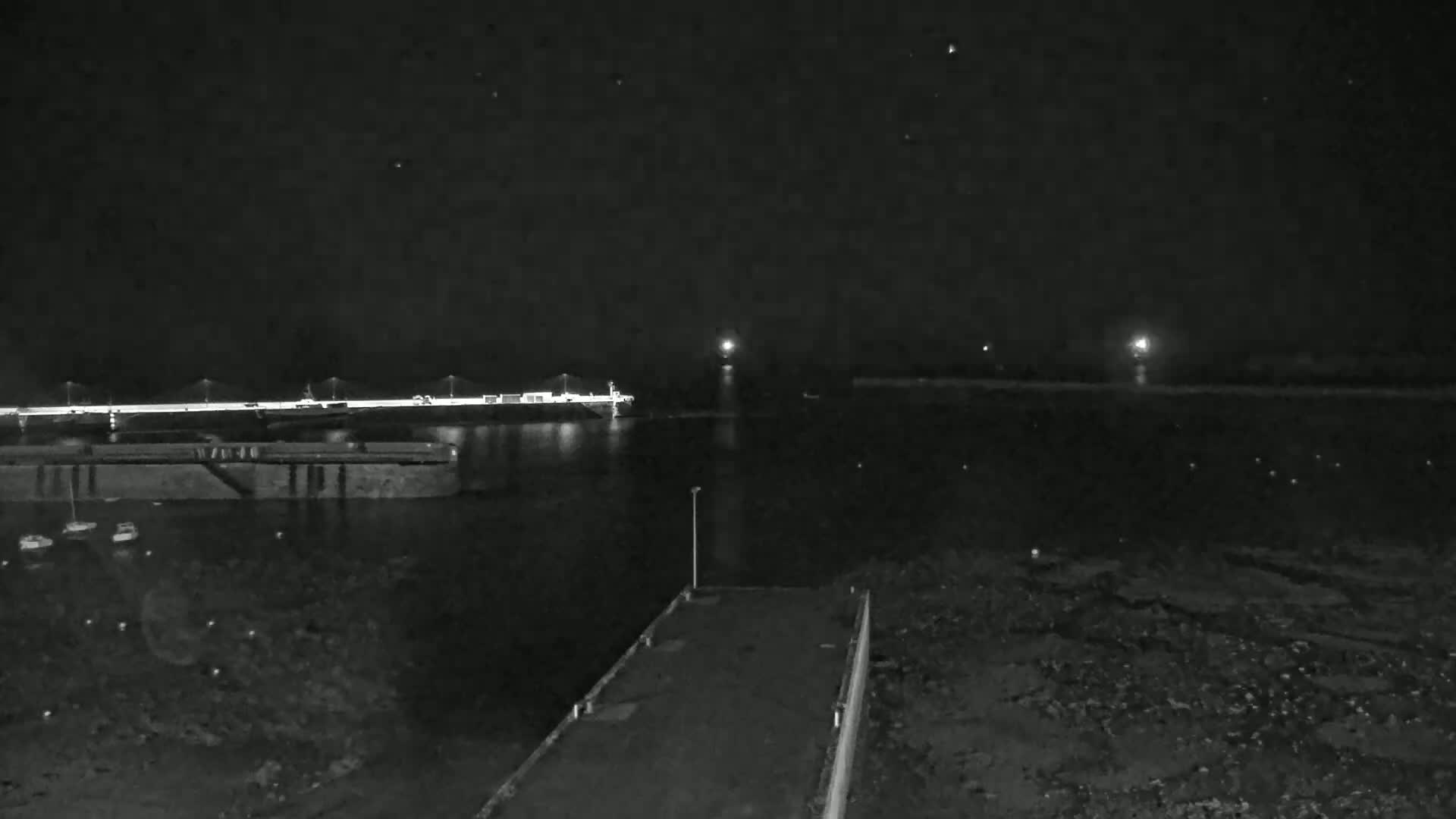 A nighttime view of a calm body of water with a bridge and docked boats in the distance, under a clear, dark sky.