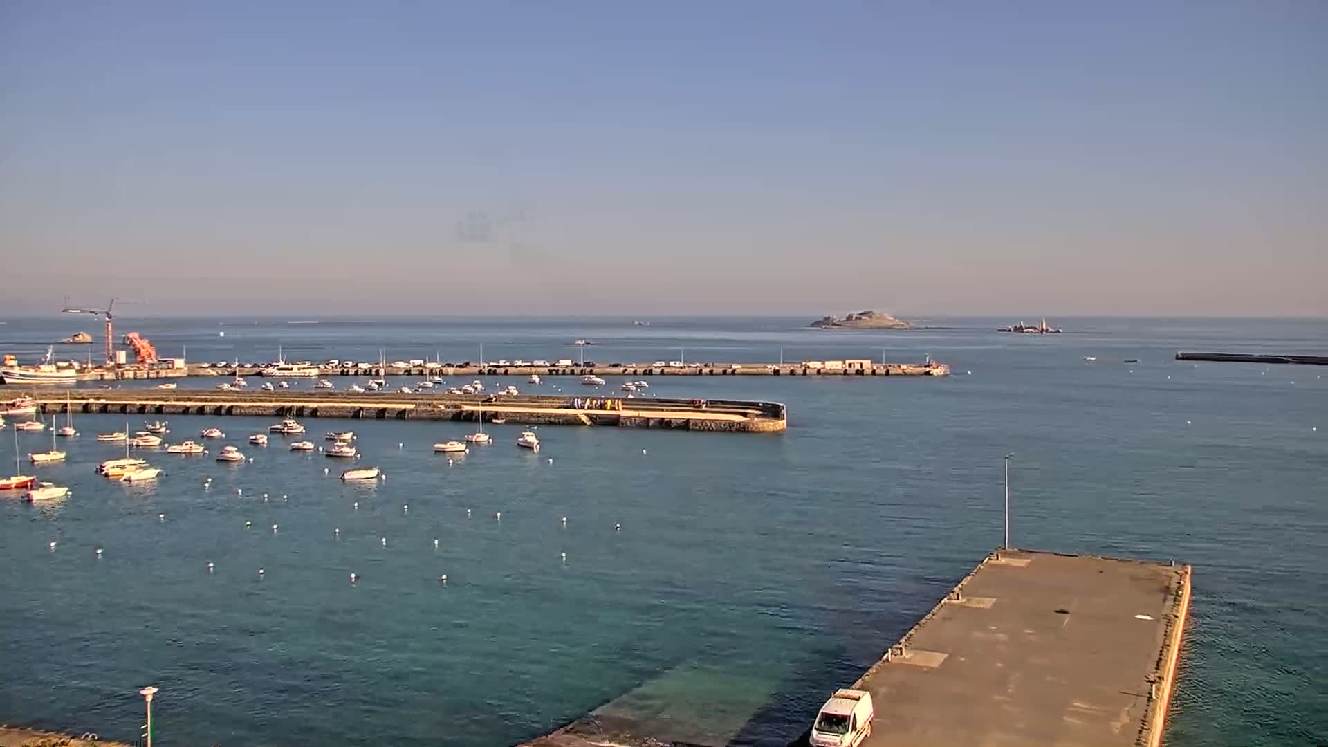 A harbor filled with numerous small boats is situated near a larger body of water under a clear, sunny sky, with a small island visible in the distance.