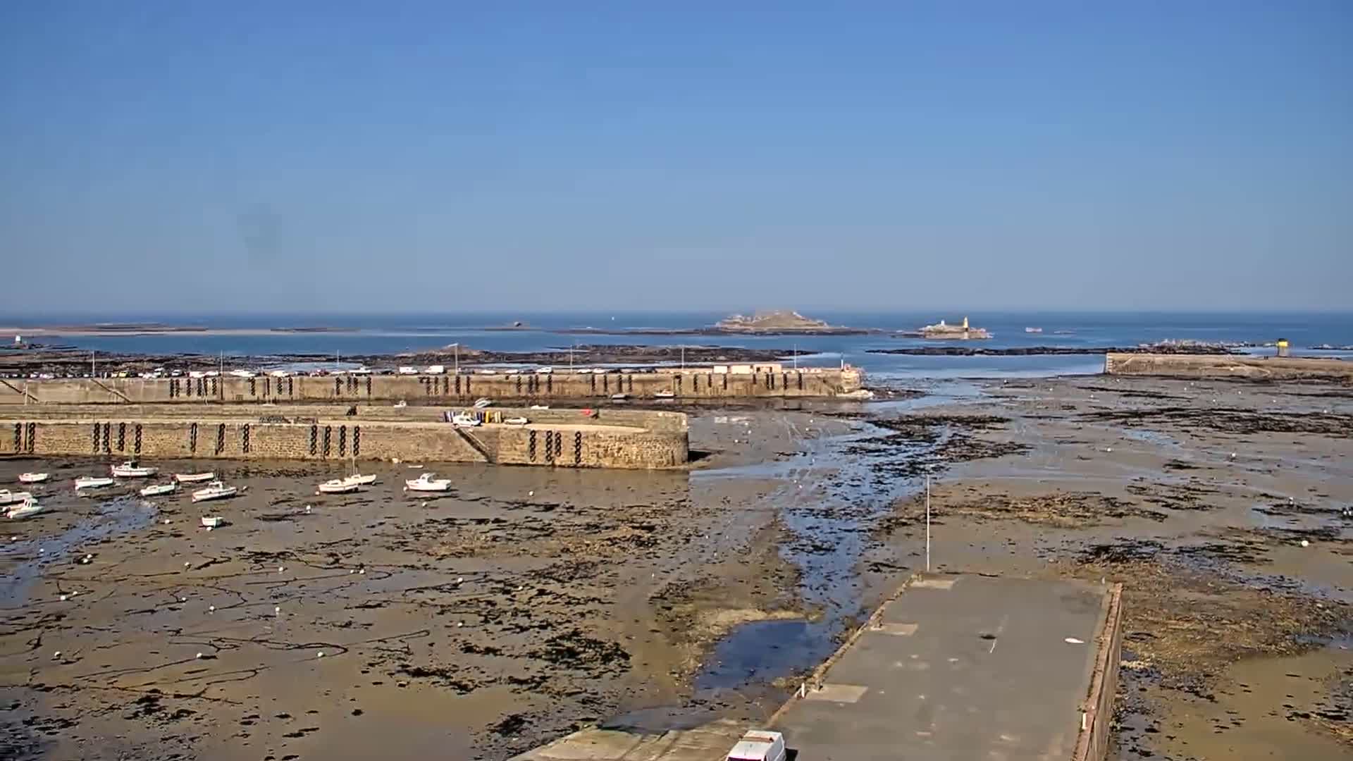 A harbor with several small boats sits at low tide on a sunny day, with an island visible in the distance.