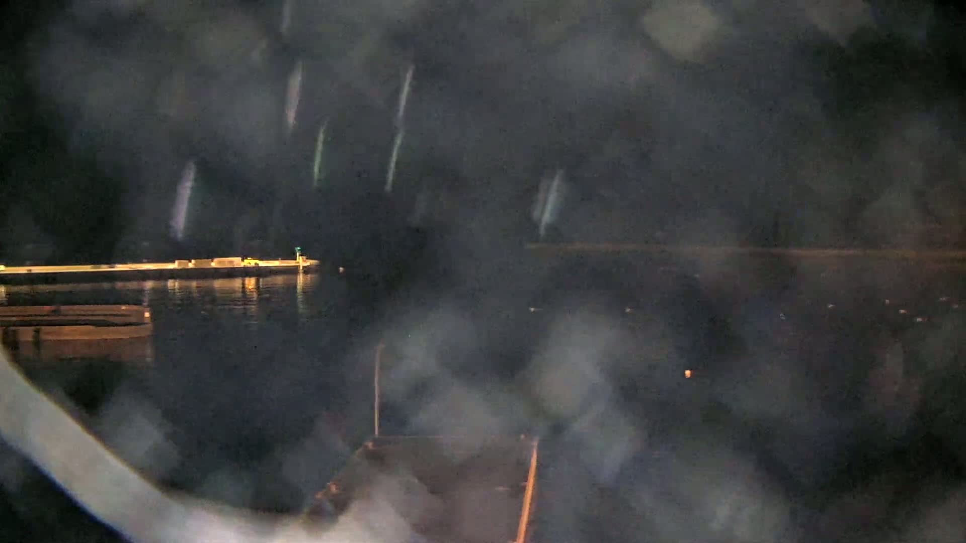 A nighttime view of a calm body of water with a dock, boats, and faint lights in the distance, seen through a hazy or rain-streaked window.