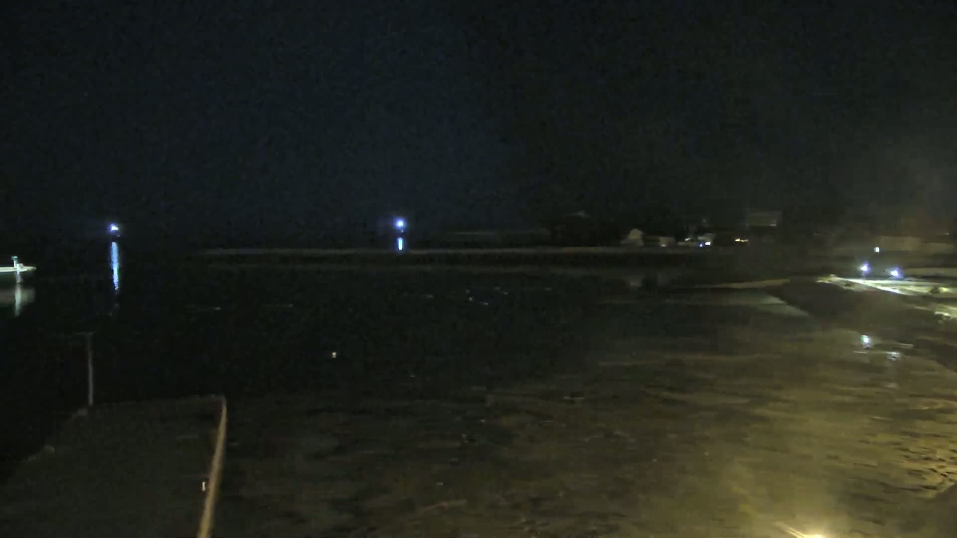 A dark, nighttime scene shows a body of water reflecting a few distant lights, with a land area illuminated by more lights in the foreground.