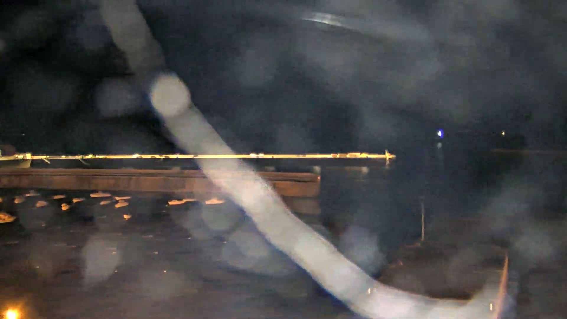 A nighttime, rain-streaked view shows a body of water with a distant illuminated pier and several small objects, possibly boats, in the water.