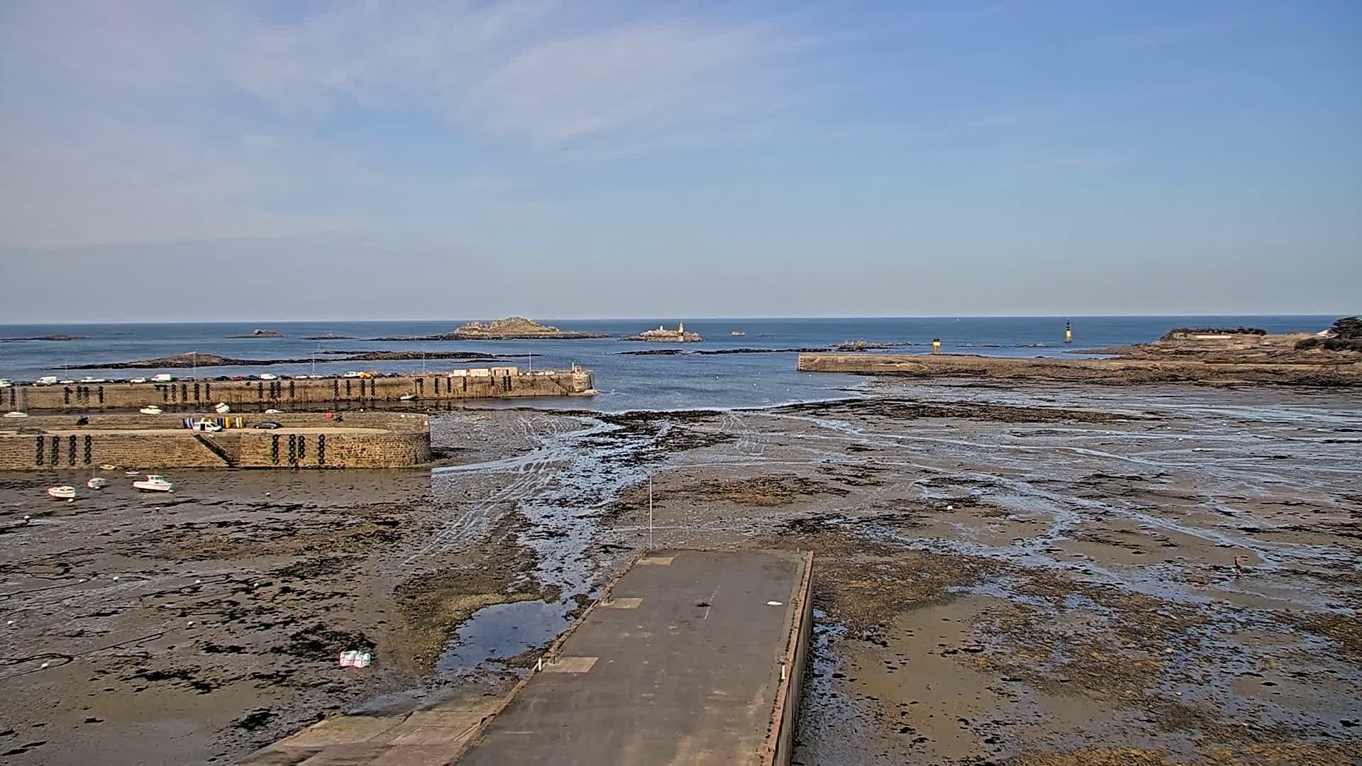 A harbor with stone walls and a few small boats is mostly exposed at low tide under a partly cloudy sky, with distant islands visible across calm water.