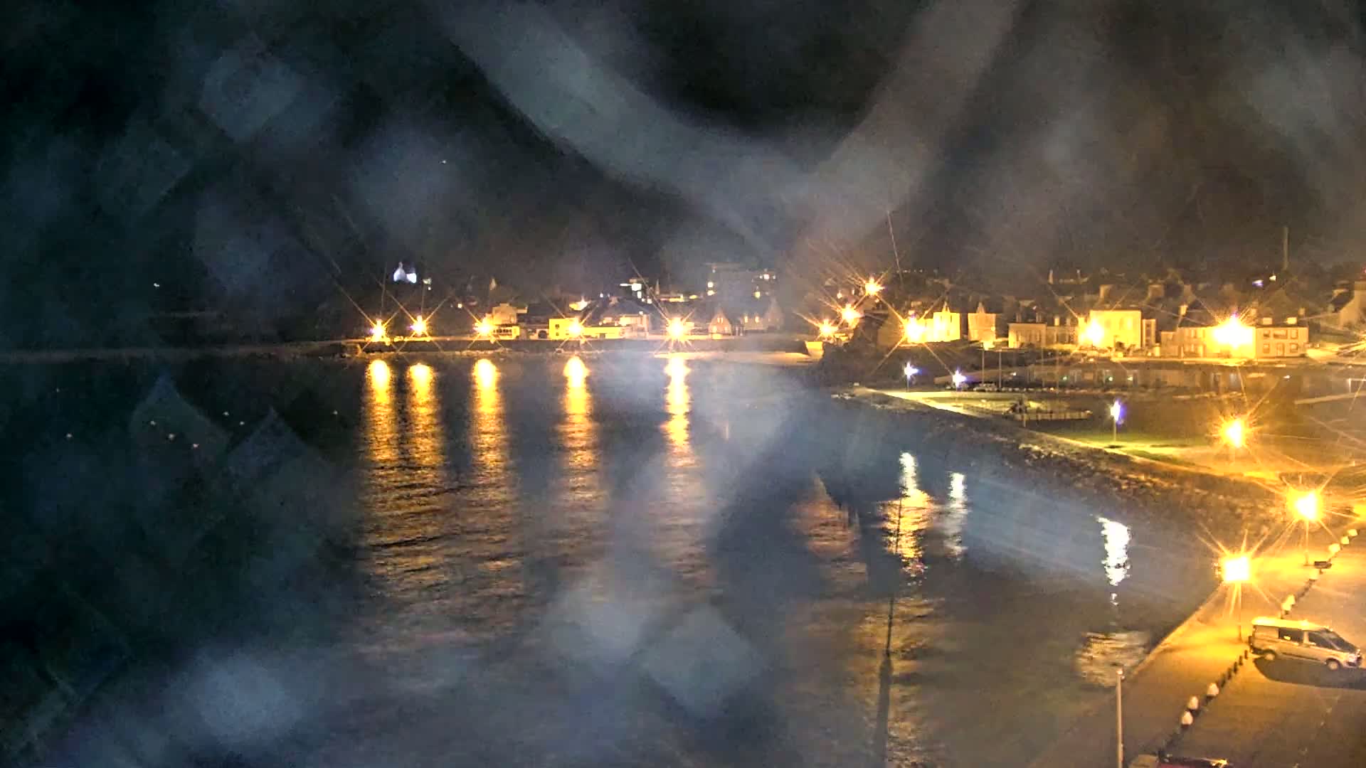 A nighttime view of a waterfront town, seen through rain-streaked glass, shows brightly lit buildings along the shore and their reflections shimmering on the dark water.