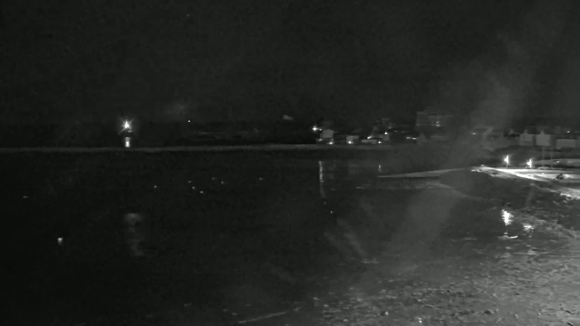 A nighttime view of a dark body of water reflecting a few distant lights, with a shoreline and buildings faintly visible in the background under a dark sky.