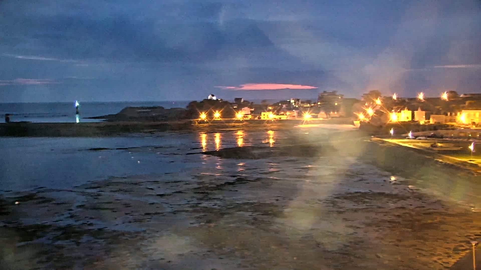 A nighttime coastal scene shows a town illuminated with lights, a calm body of water reflecting the lights, and a distant lighthouse under a twilight sky.