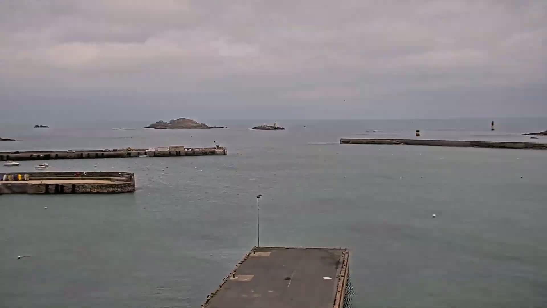 A harbor with several docks and small boats, under an overcast sky, is seen with a small island and a lighthouse visible in the distance.