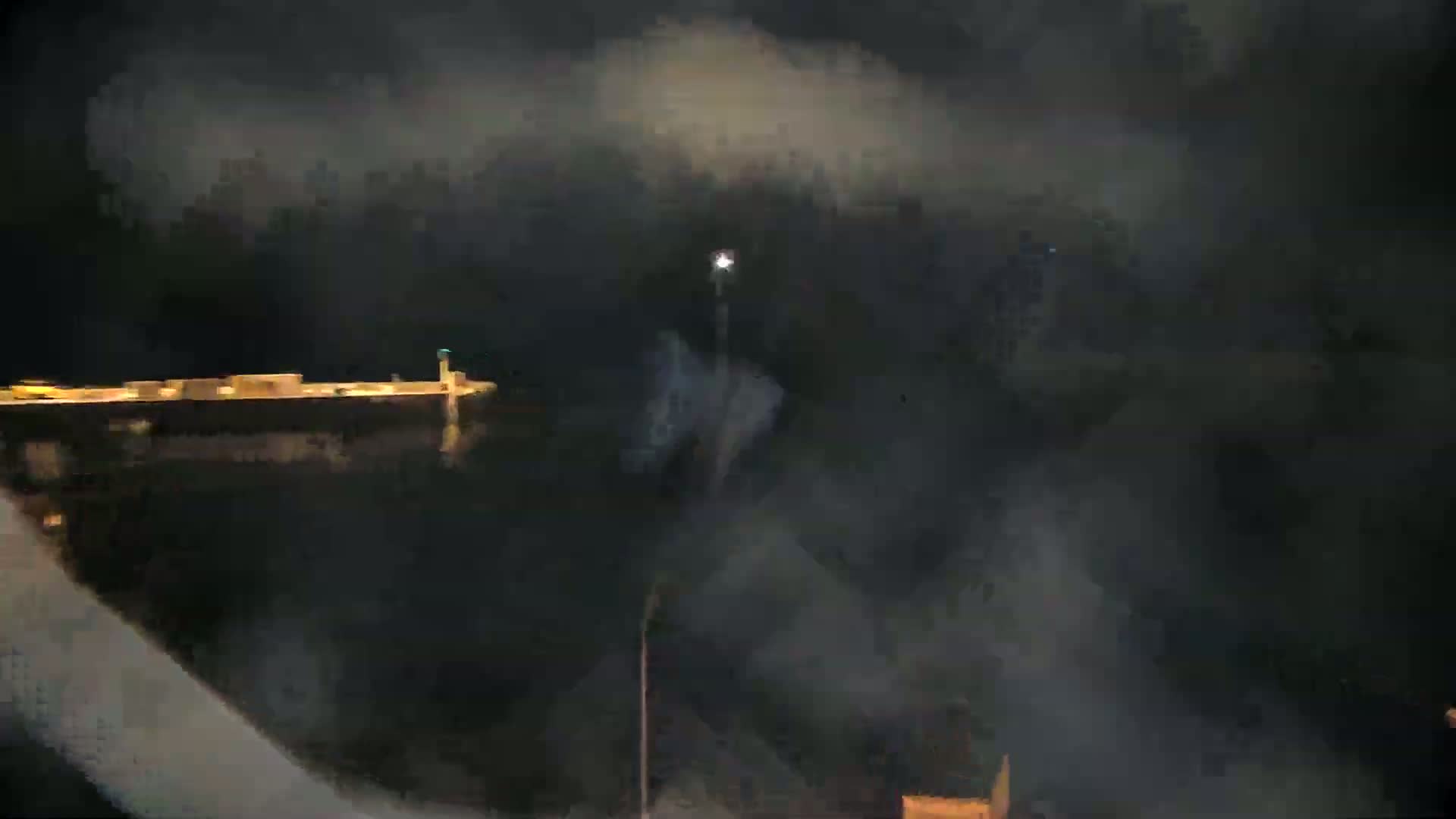 A dark, foggy night scene shows a long, illuminated vessel docked at a pier, with a bright light visible in the distance.