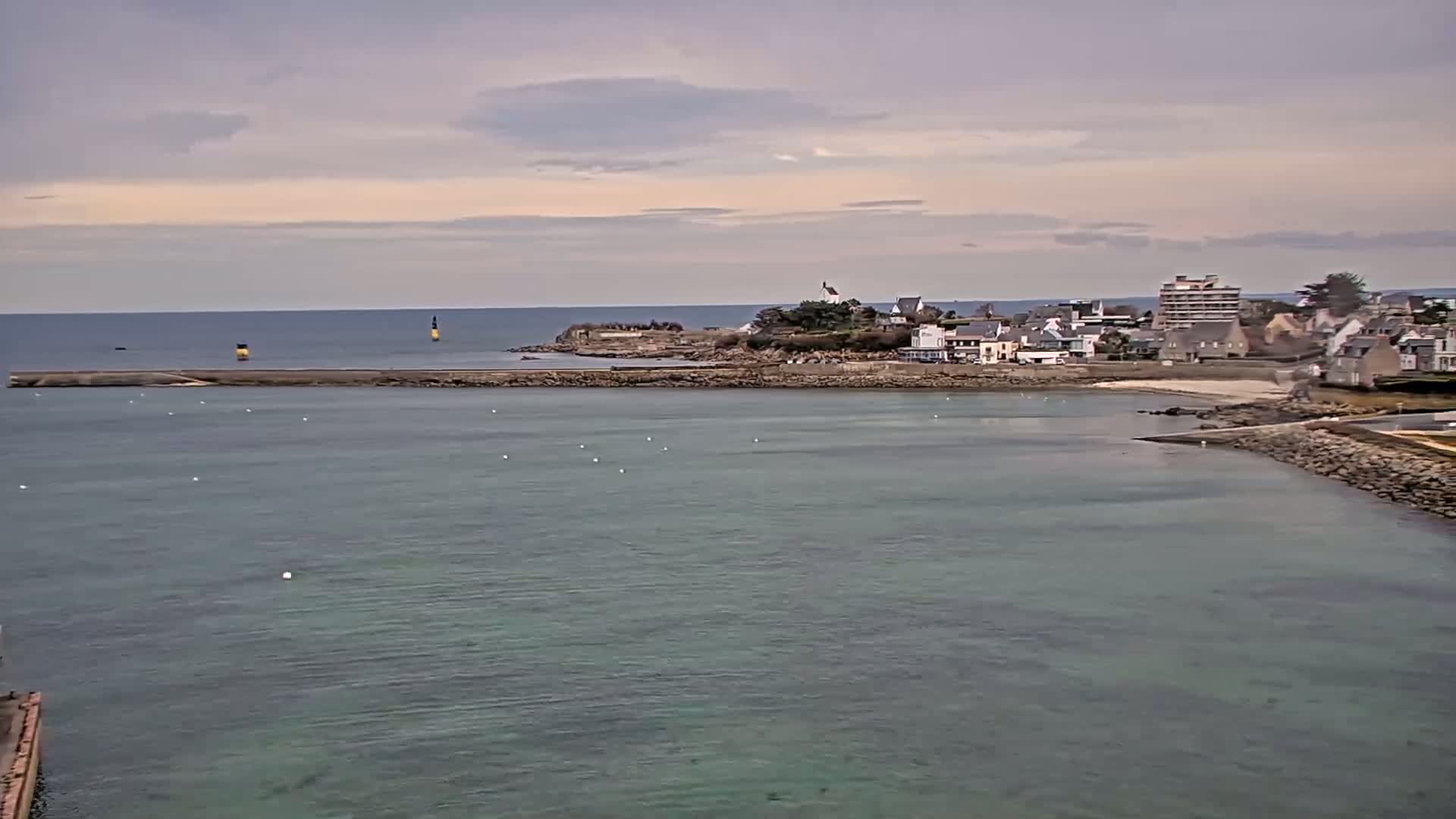 A coastal town with a small beach sits on a rocky point under a mostly cloudy sky, with calm, shallow water stretching out to the horizon.