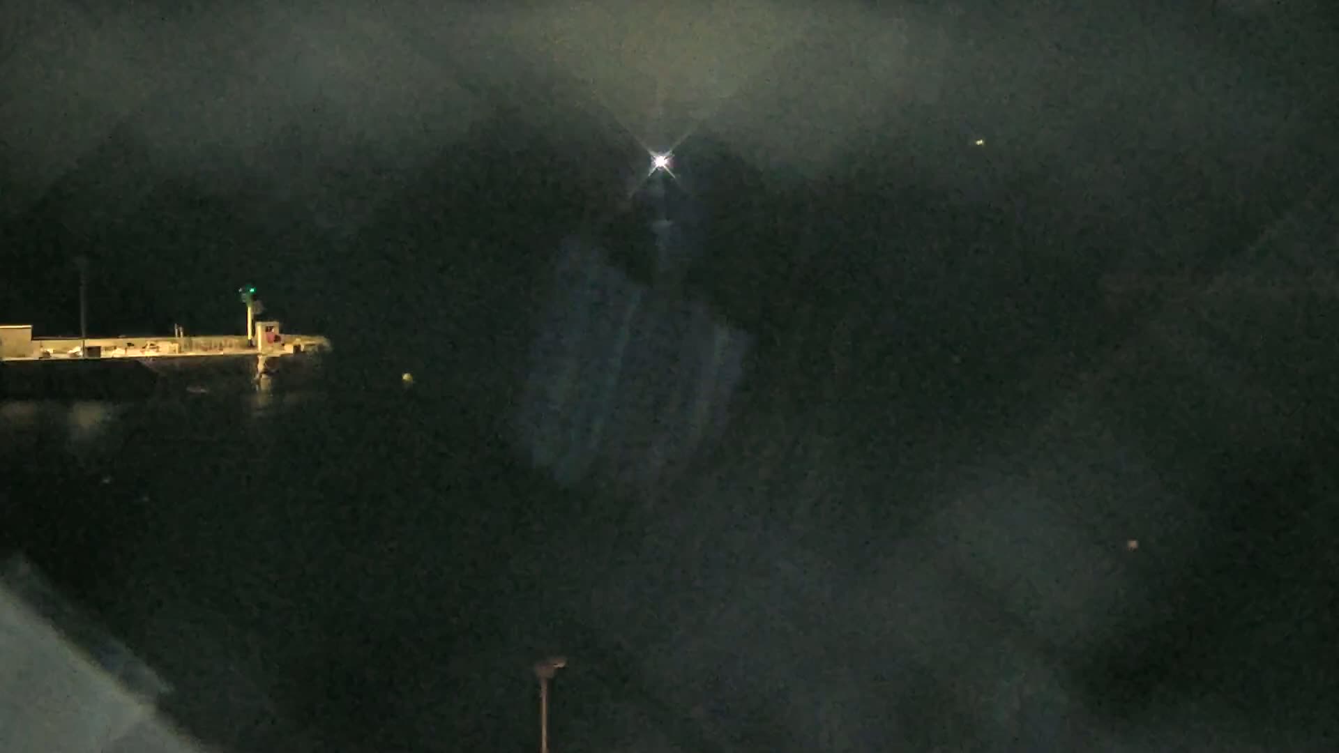 A dark, foggy night scene shows a distant structure with a light on a pier, illuminated by a bright light source in the upper center.