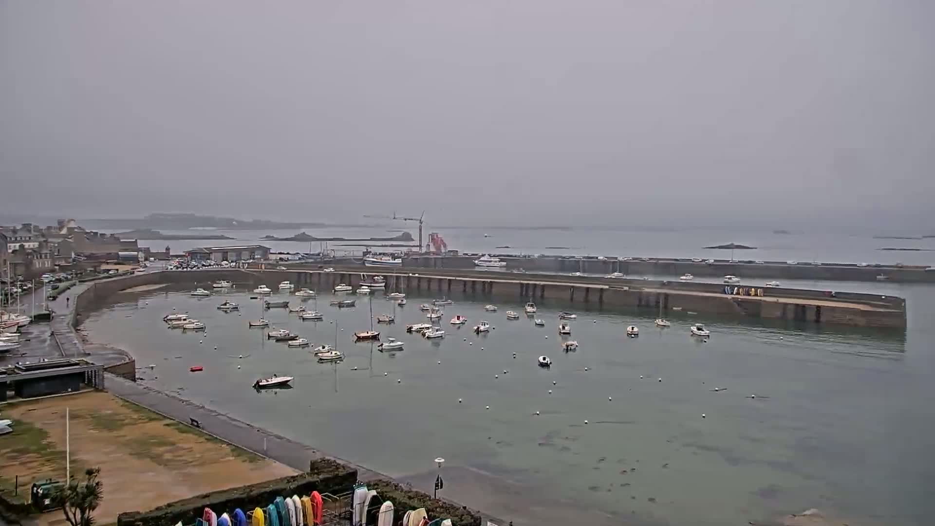 A harbor filled with numerous small boats is seen on an overcast day, with a town and distant coastline visible in the background.