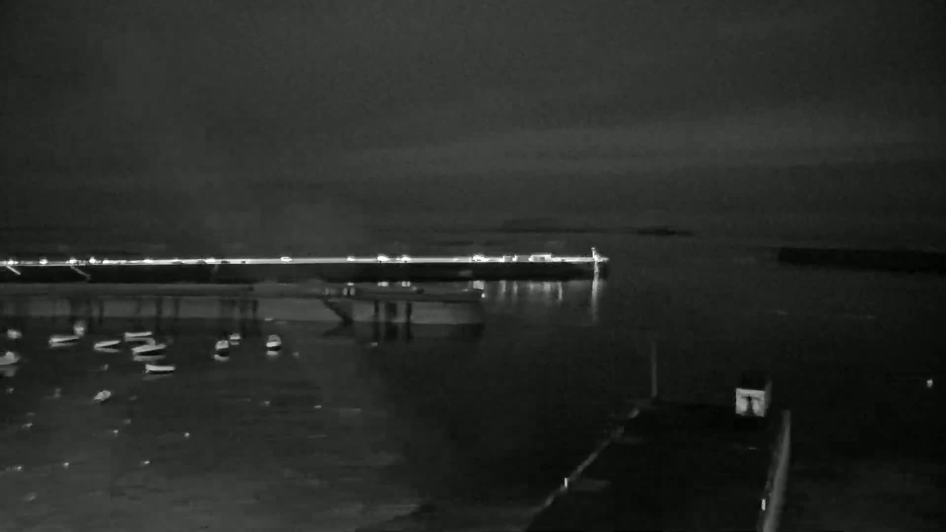 A nighttime, low-light view of a harbor with numerous small boats at rest, a pier with lights in the distance, and calm water.