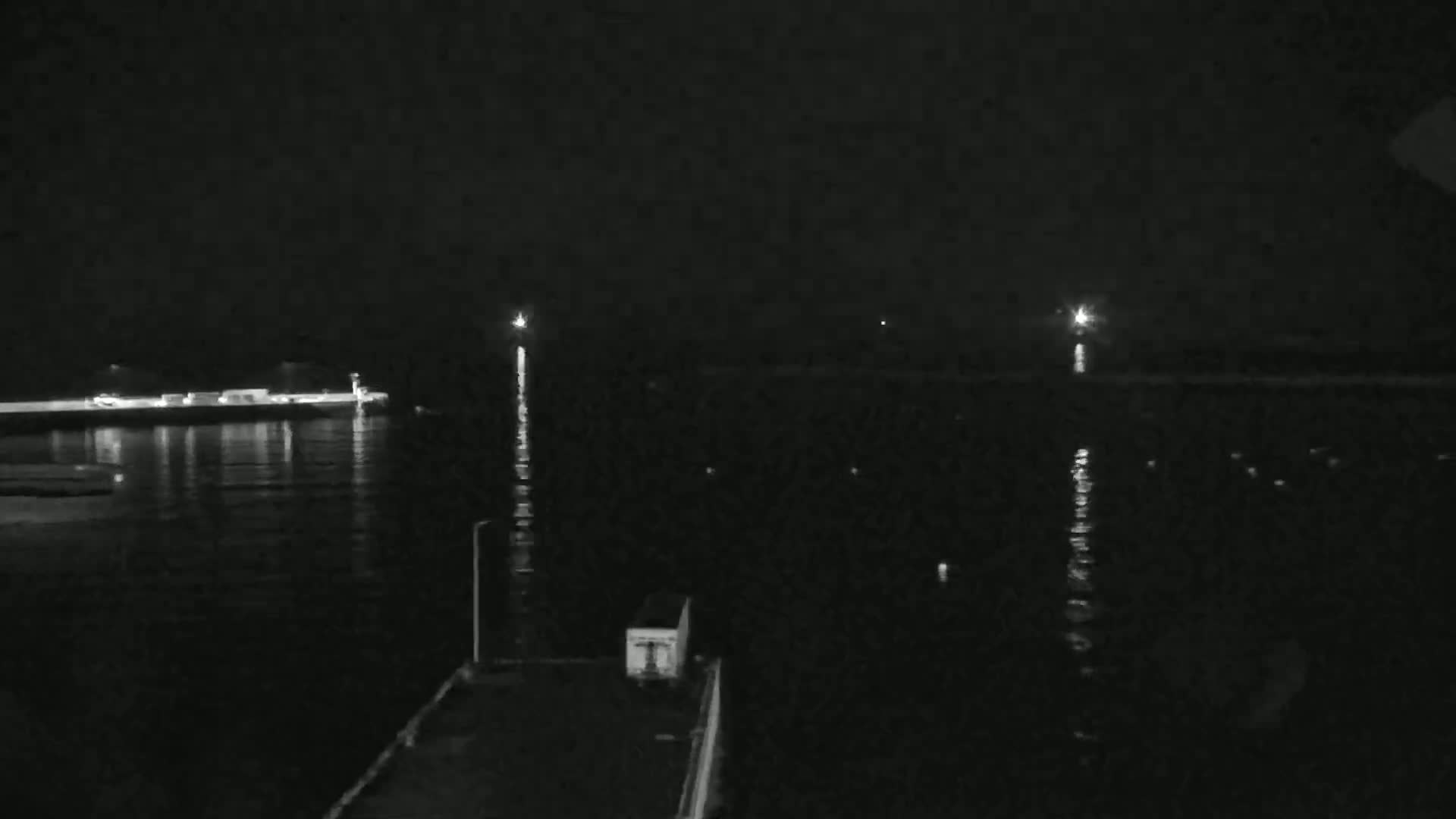 A nighttime view of calm water reflecting lights from a distant shore and two beacons, with a dark structure in the foreground.