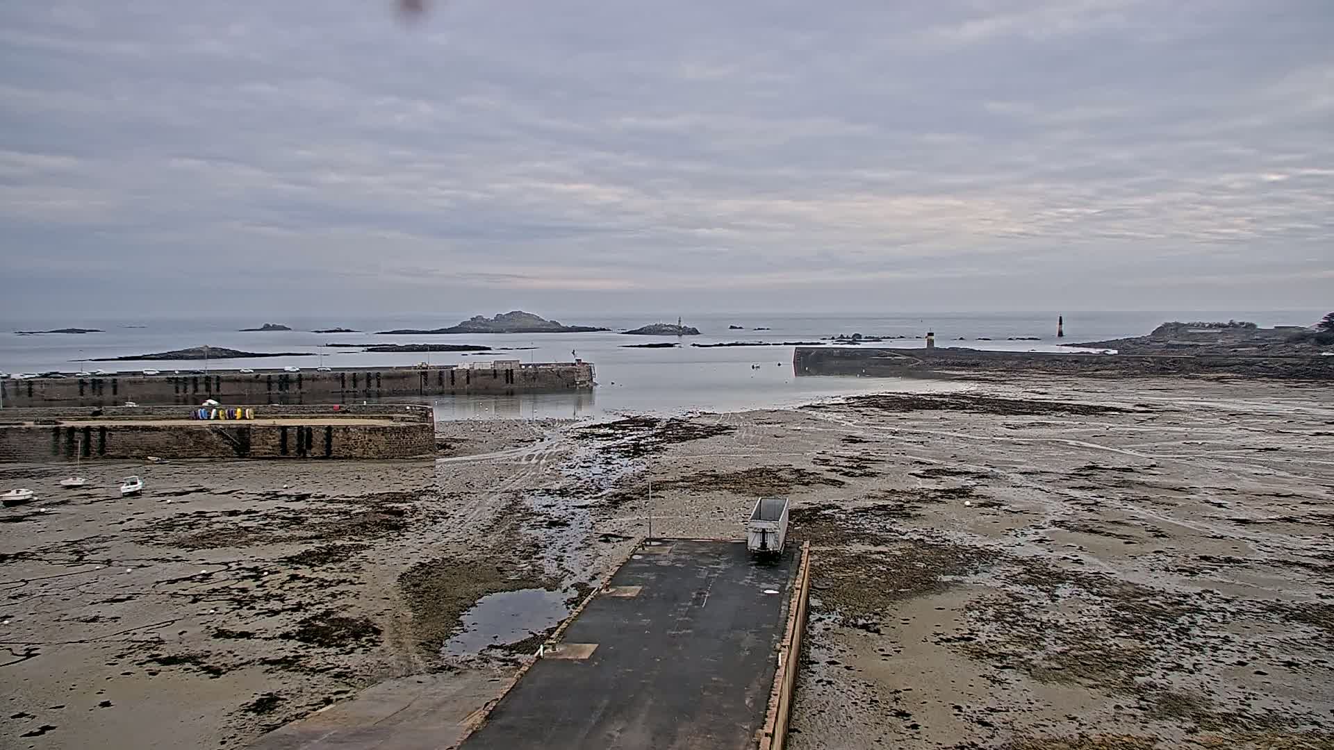 A harbor with a mostly exposed, mud-flat seabed under an overcast sky shows a few small boats near stone harbor walls, and a distant island with a lighthouse.