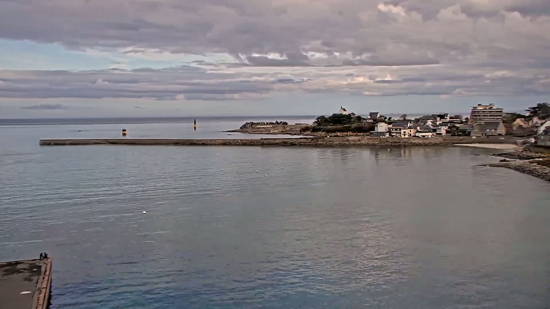 A calm body of water under a cloudy sky shows a small coastal town with buildings and a low seawall extending into the water.