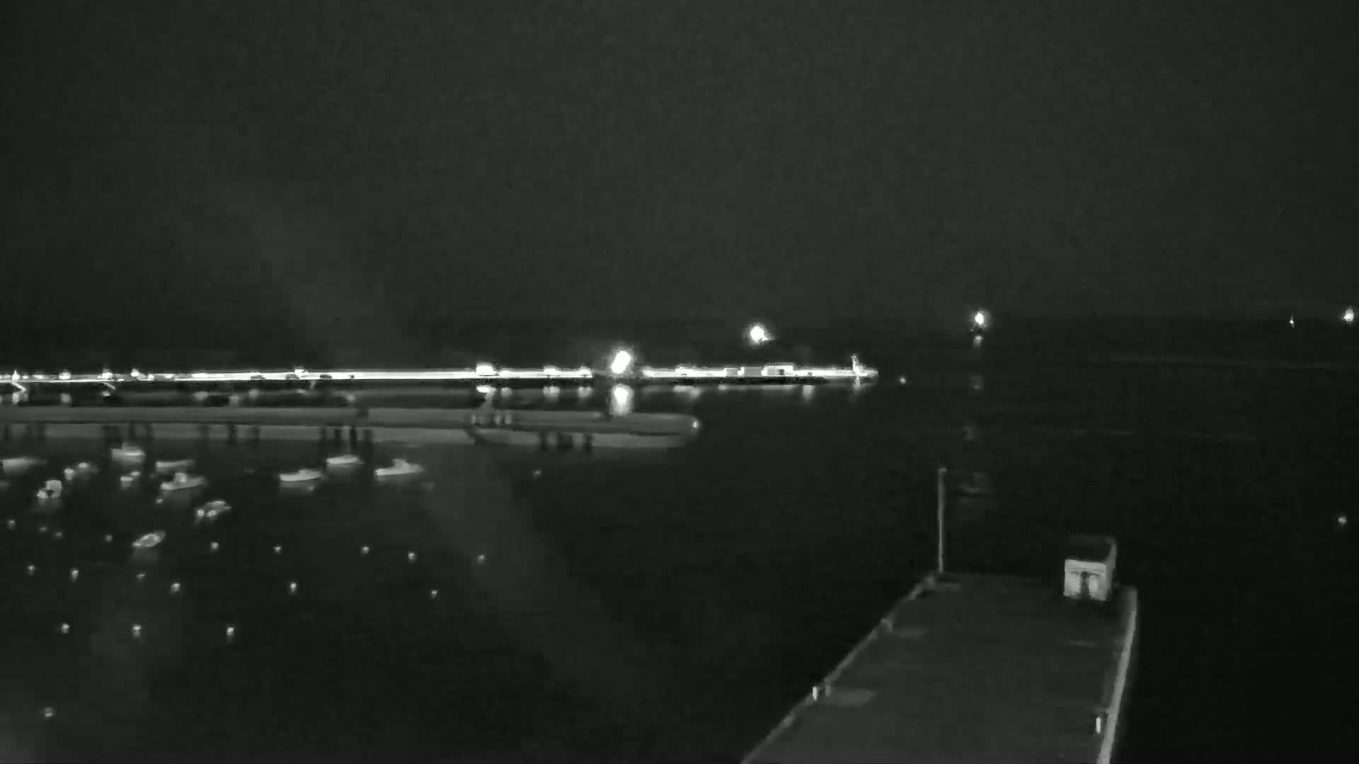 A nighttime view of a harbor with numerous small boats moored in the water, a lit shoreline in the distance, and a dark barge or dock in the foreground under a dark sky.