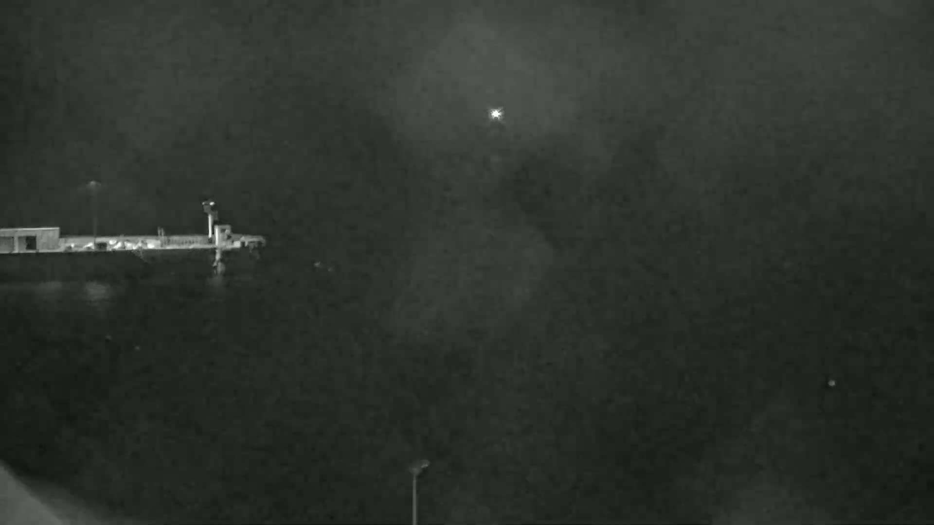 A nighttime, low-light image shows a dark body of water with a dock and a structure on the left, and a bright light in the distance.