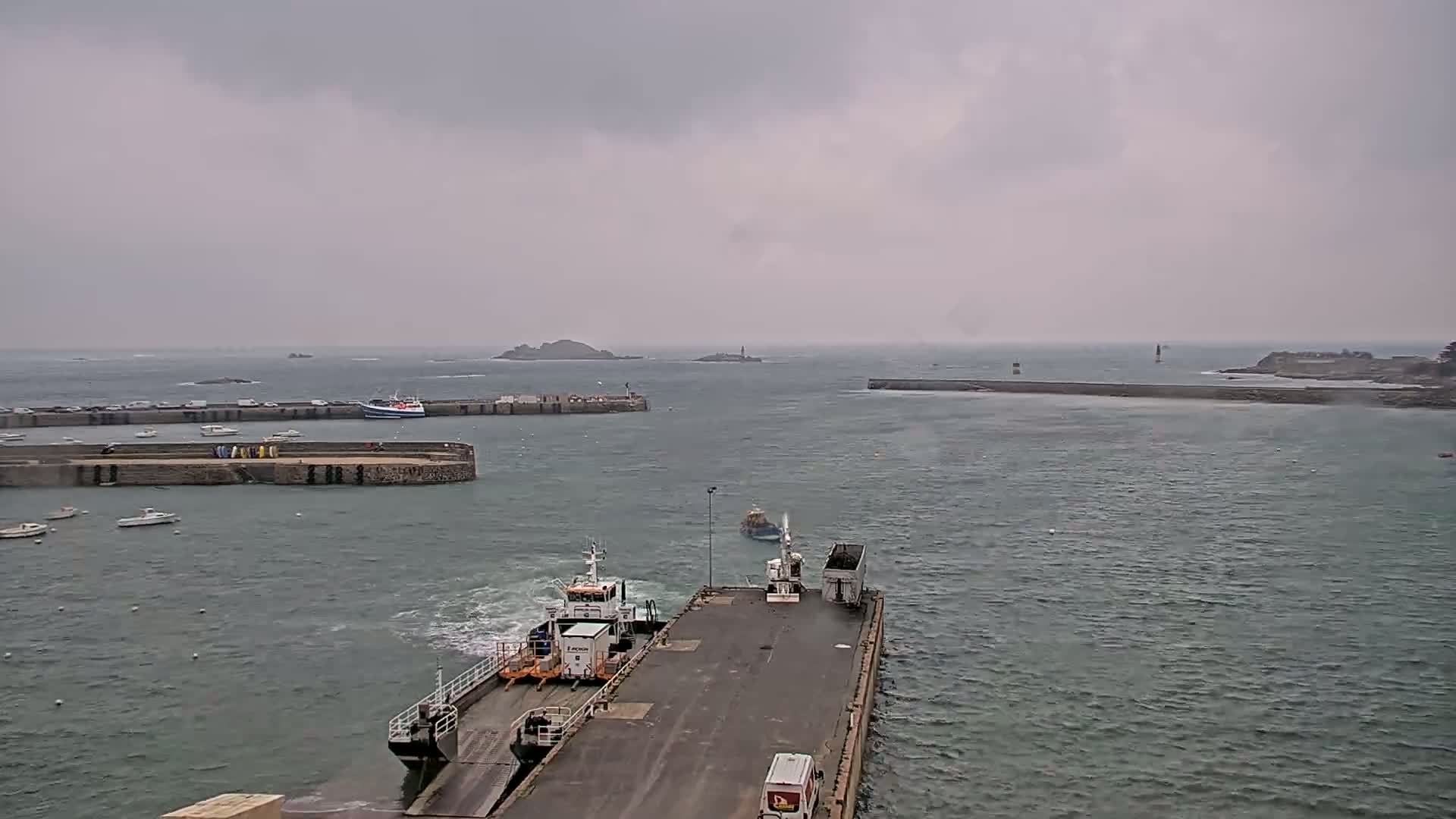 A ferry approaches a dock in a harbor on an overcast day, with several small boats and islands in the distance.