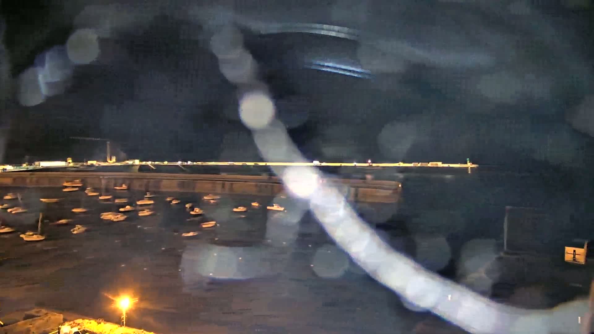 Numerous small boats are moored in a harbor at night, viewed through rain-covered glass.