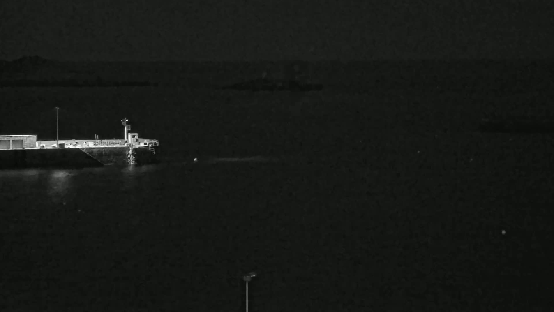 A large dark vessel is docked at a pier at night, with calm water surrounding it under a dark sky.