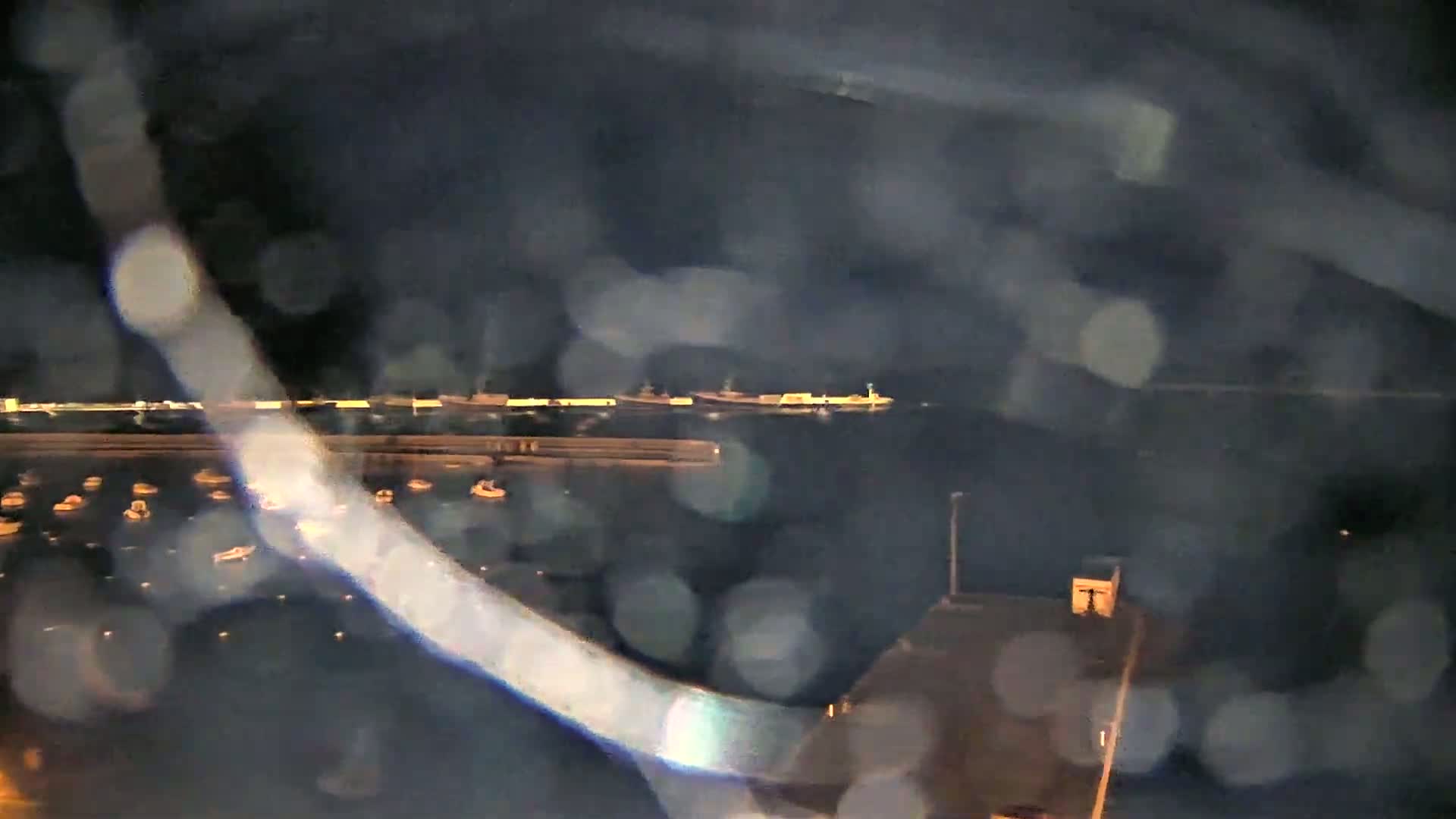 A nighttime, rain-blurred view shows a harbor with several small boats and larger vessels moored at a dock.