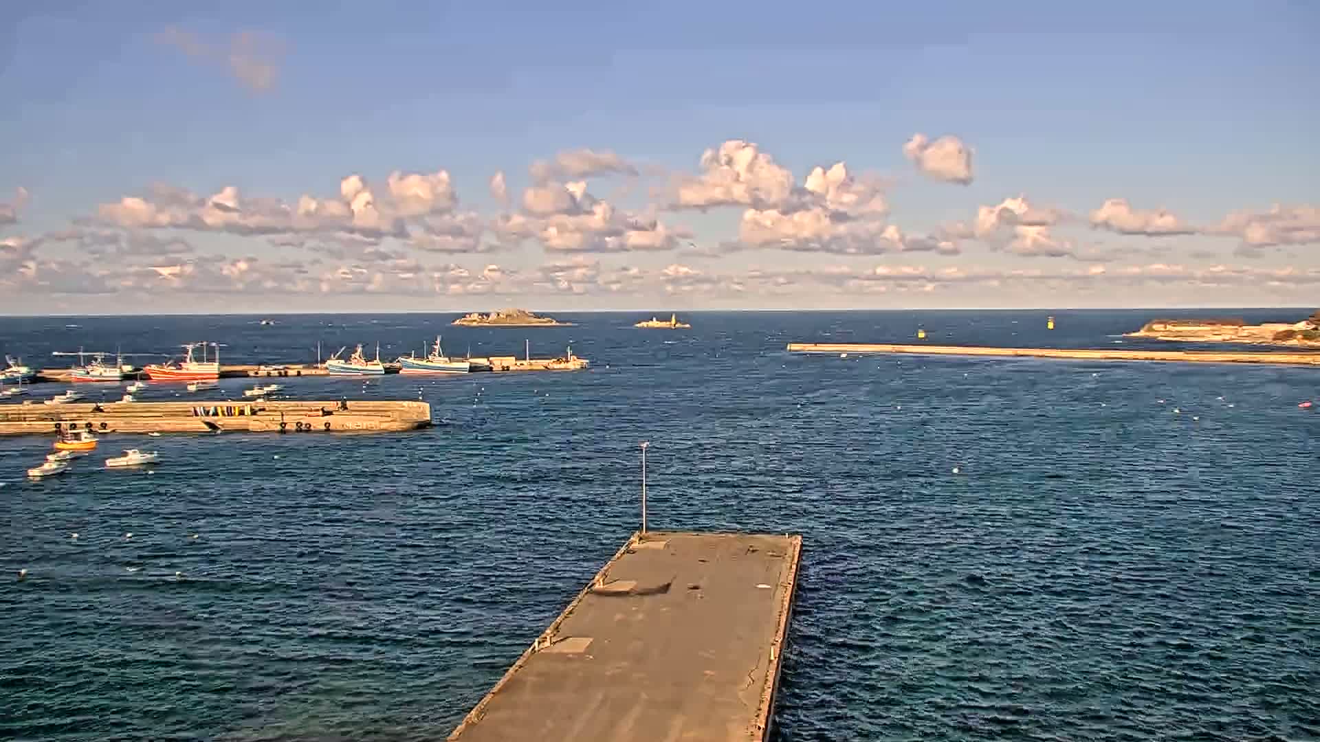 A harbor filled with fishing boats is seen under a partly cloudy sky, with calm water and distant islands visible on the horizon.