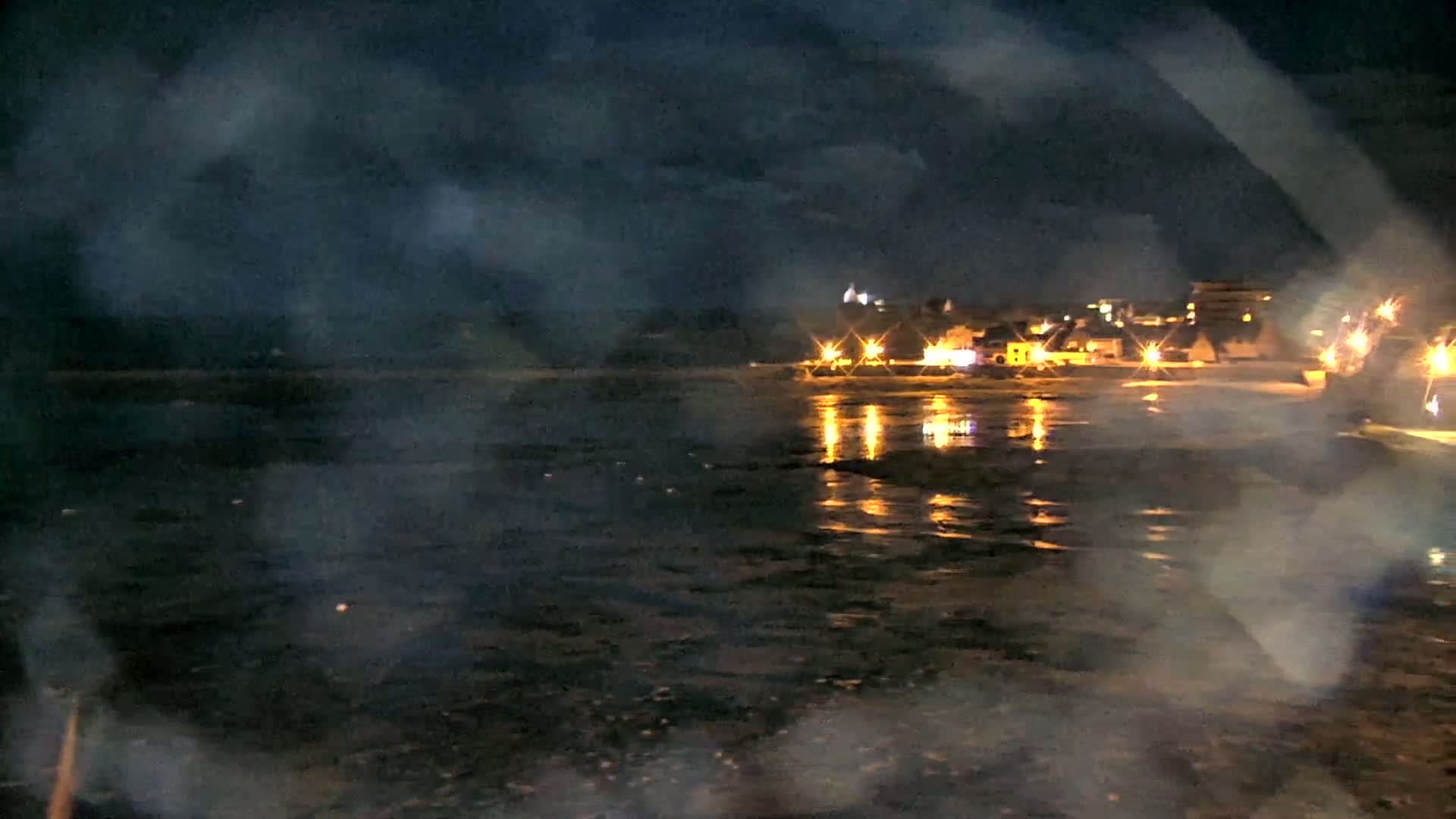 A nighttime view of a dark, possibly wet, shoreline with a town's lights reflecting on the water under a cloudy sky.