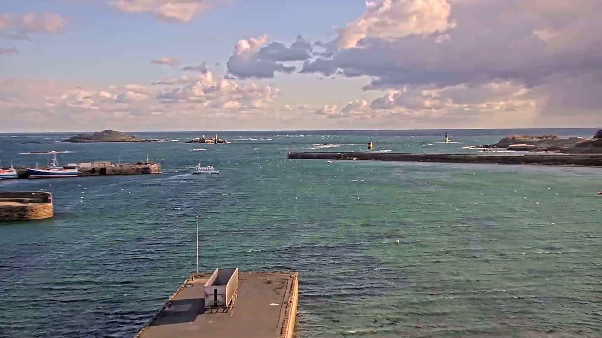 A harbor with several boats is seen under a partly cloudy sky, with a small island visible in the distance.
