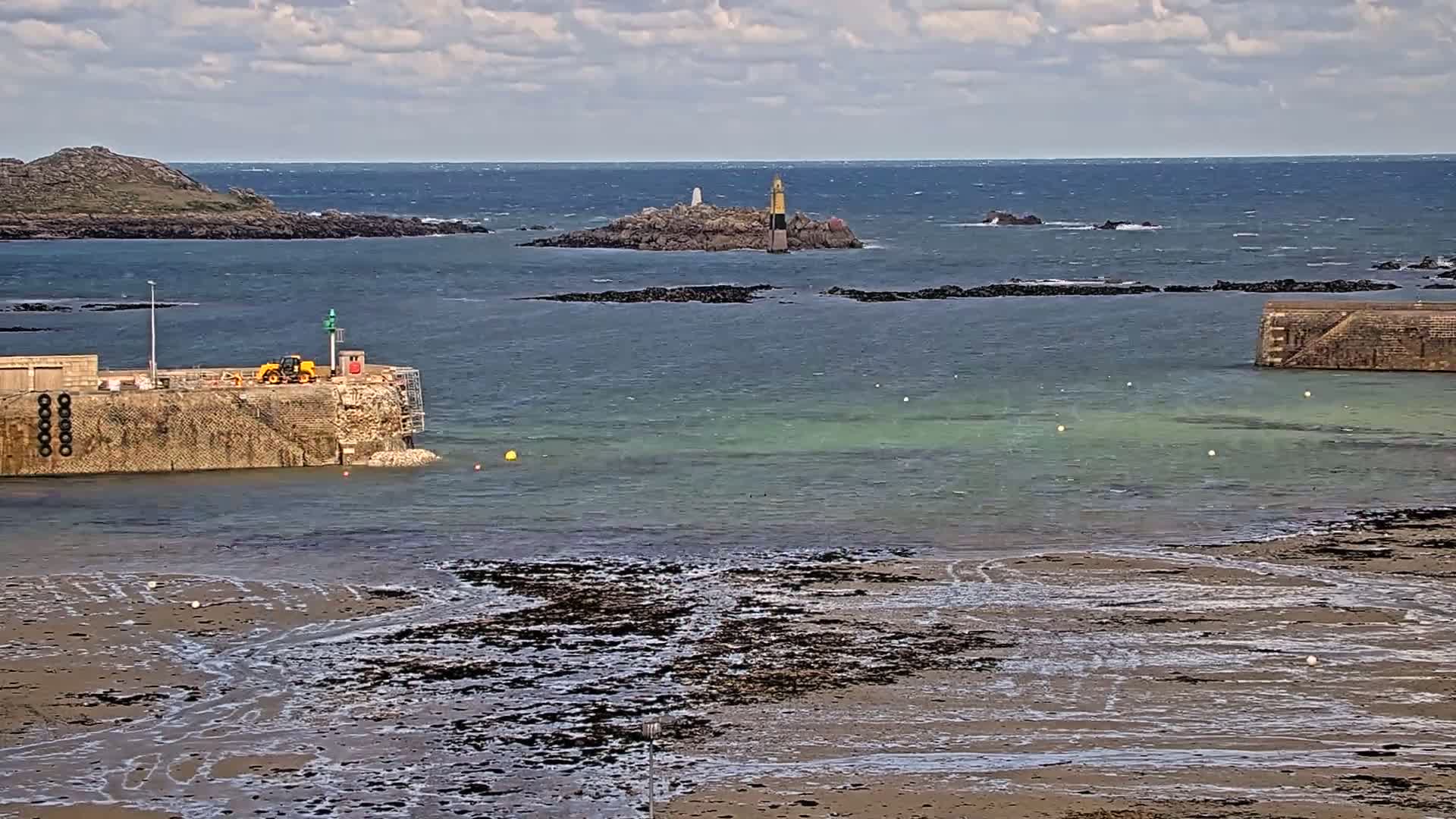 A partially exposed sandy shore leads to a calm, shallow harbor with a small island featuring a lighthouse in the distance under a partly cloudy sky.