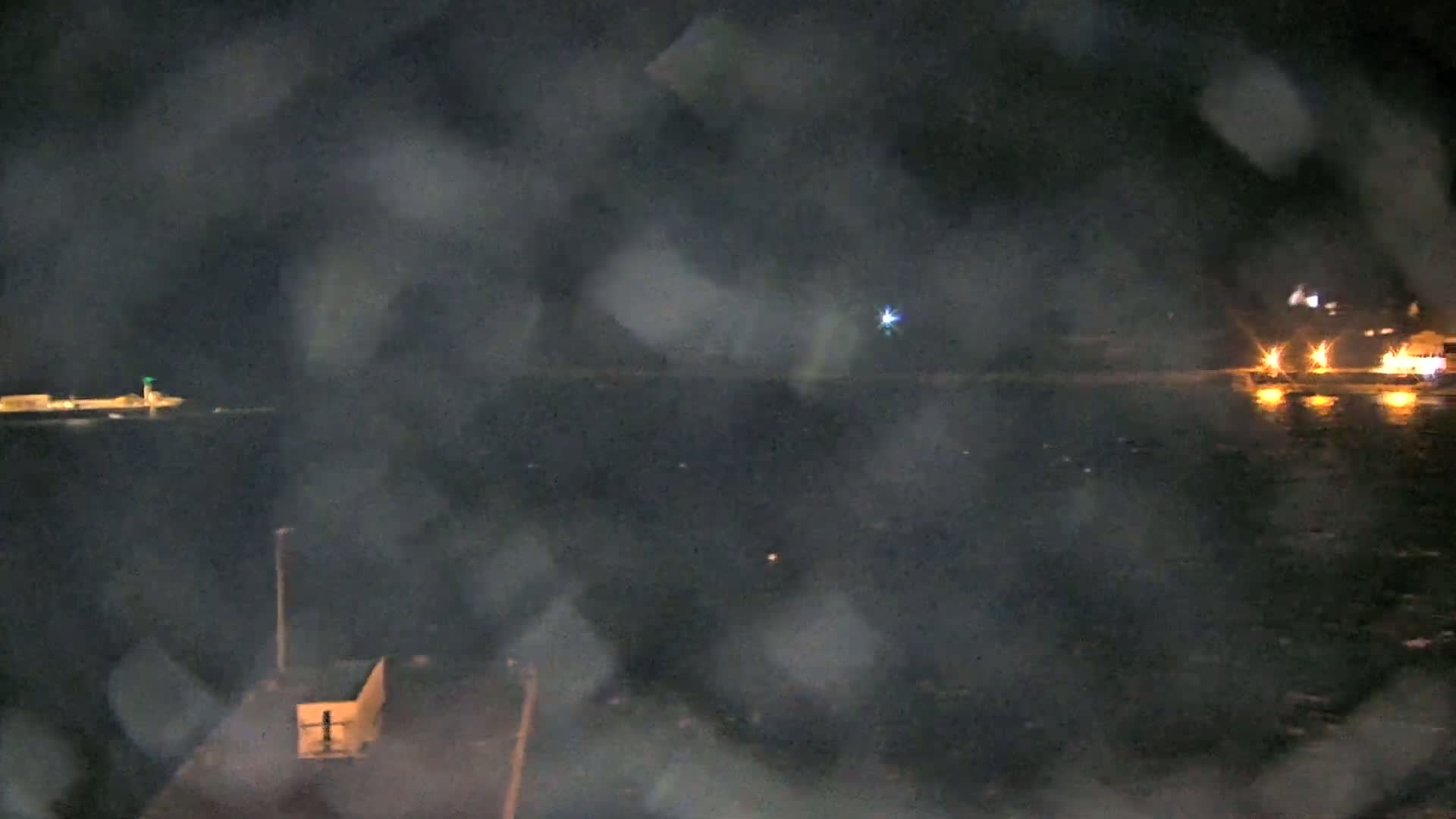 A nighttime view of a dark body of water with blurry lights on the far shore and a small structure in the foreground, possibly viewed through rain or fog.