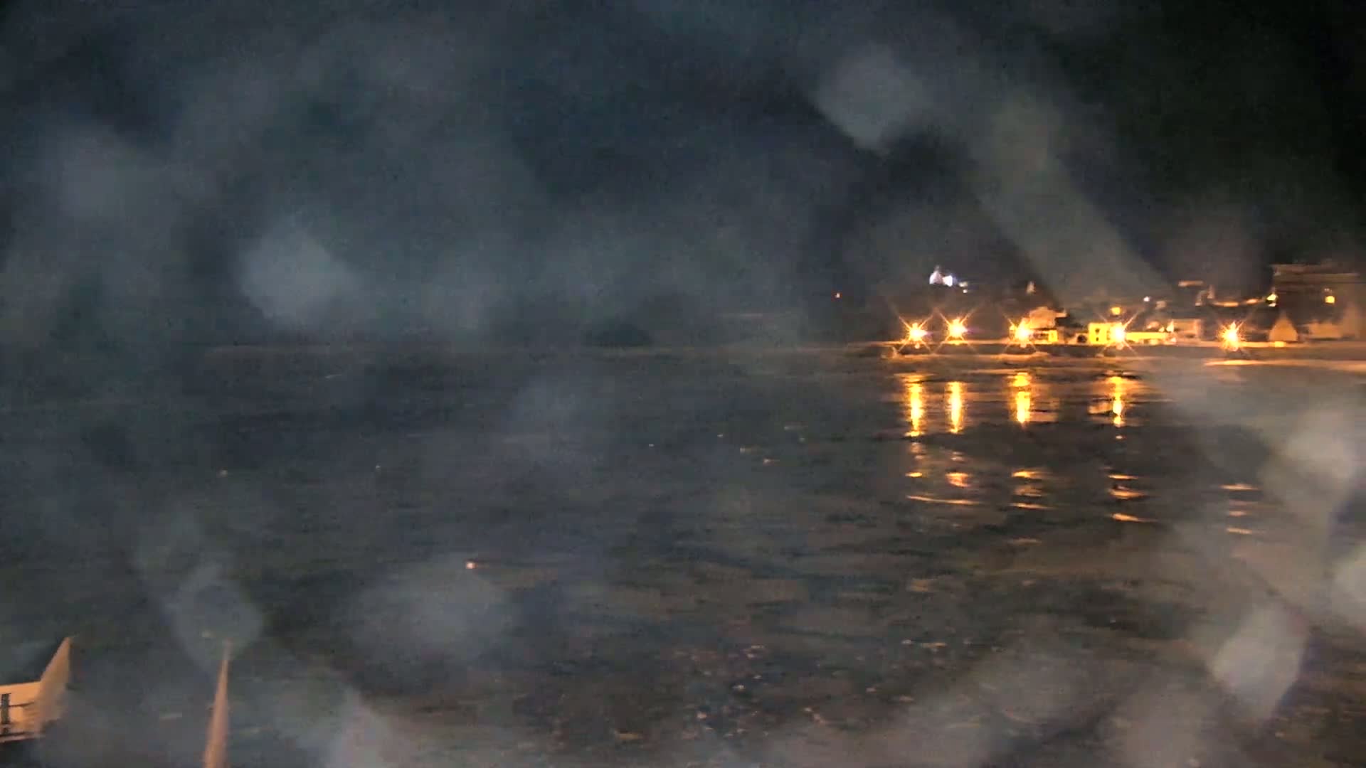 A nighttime view of a calm body of water reflecting the lights of a distant town, seen through a rain-streaked window.