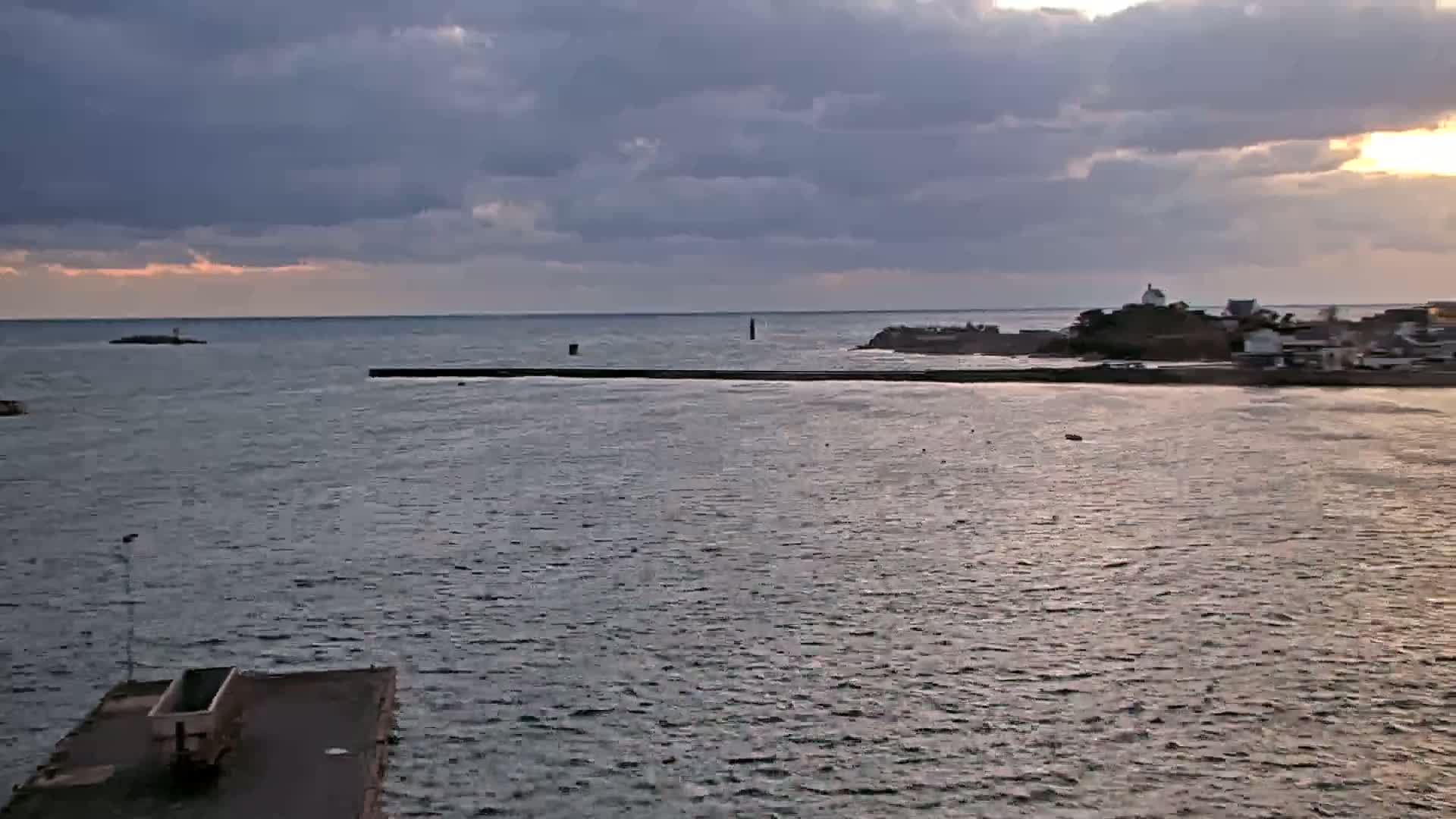 A calm body of water under a partly cloudy sunset sky shows a small town and breakwater in the distance.