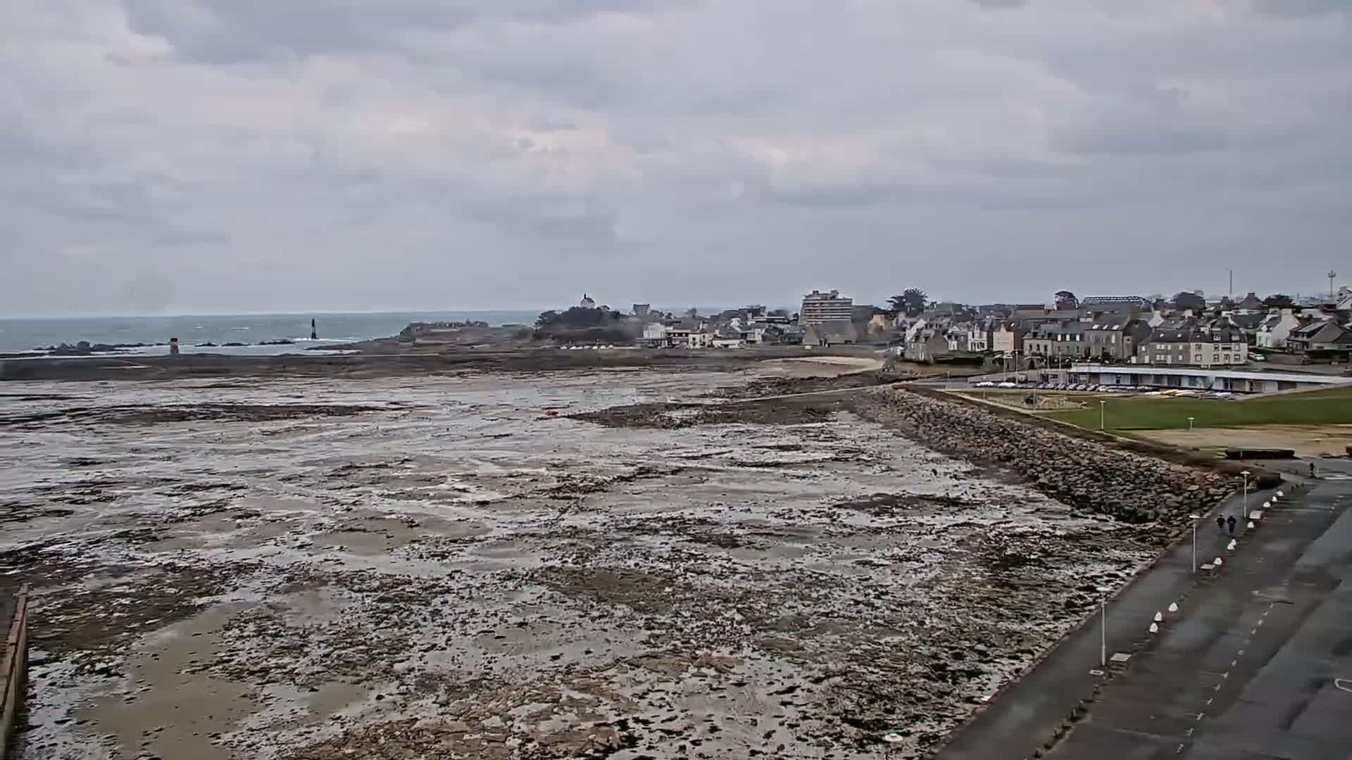 A wide shot shows a coastal town under an overcast sky, with a large expanse of exposed tidal flats stretching between the town and the ocean.