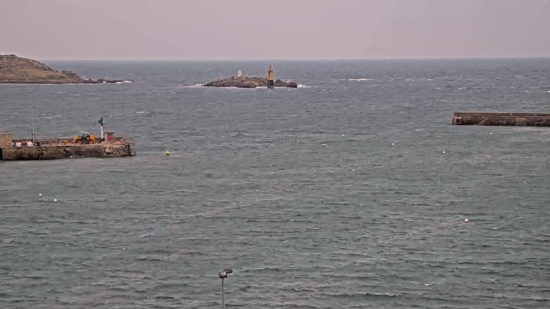 A small island with a lighthouse is visible in a choppy sea, near a harbor with a small piece of construction equipment on a pier.