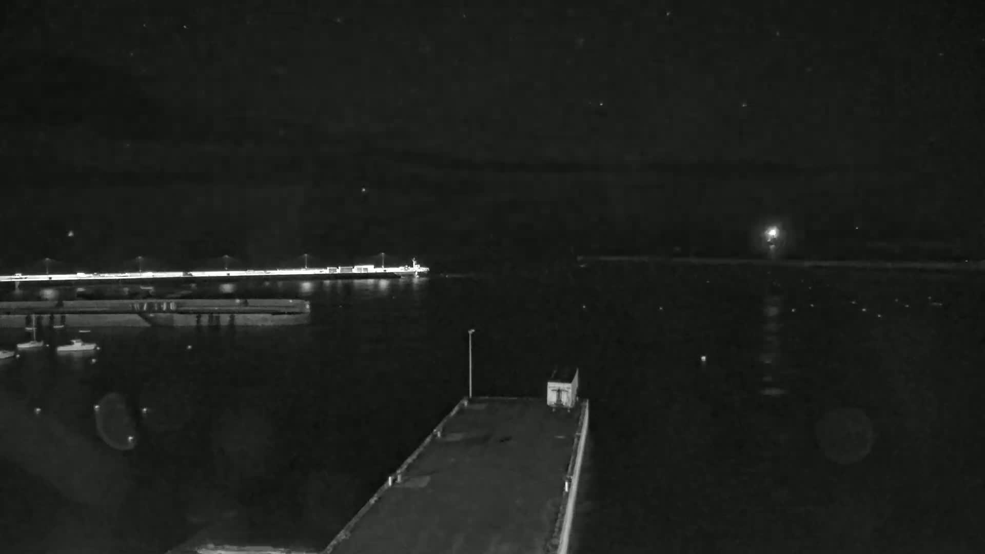 A nighttime view of a harbor shows a pier with a small structure, boats in the water, and a distant, illuminated shoreline under a clear, starry sky.