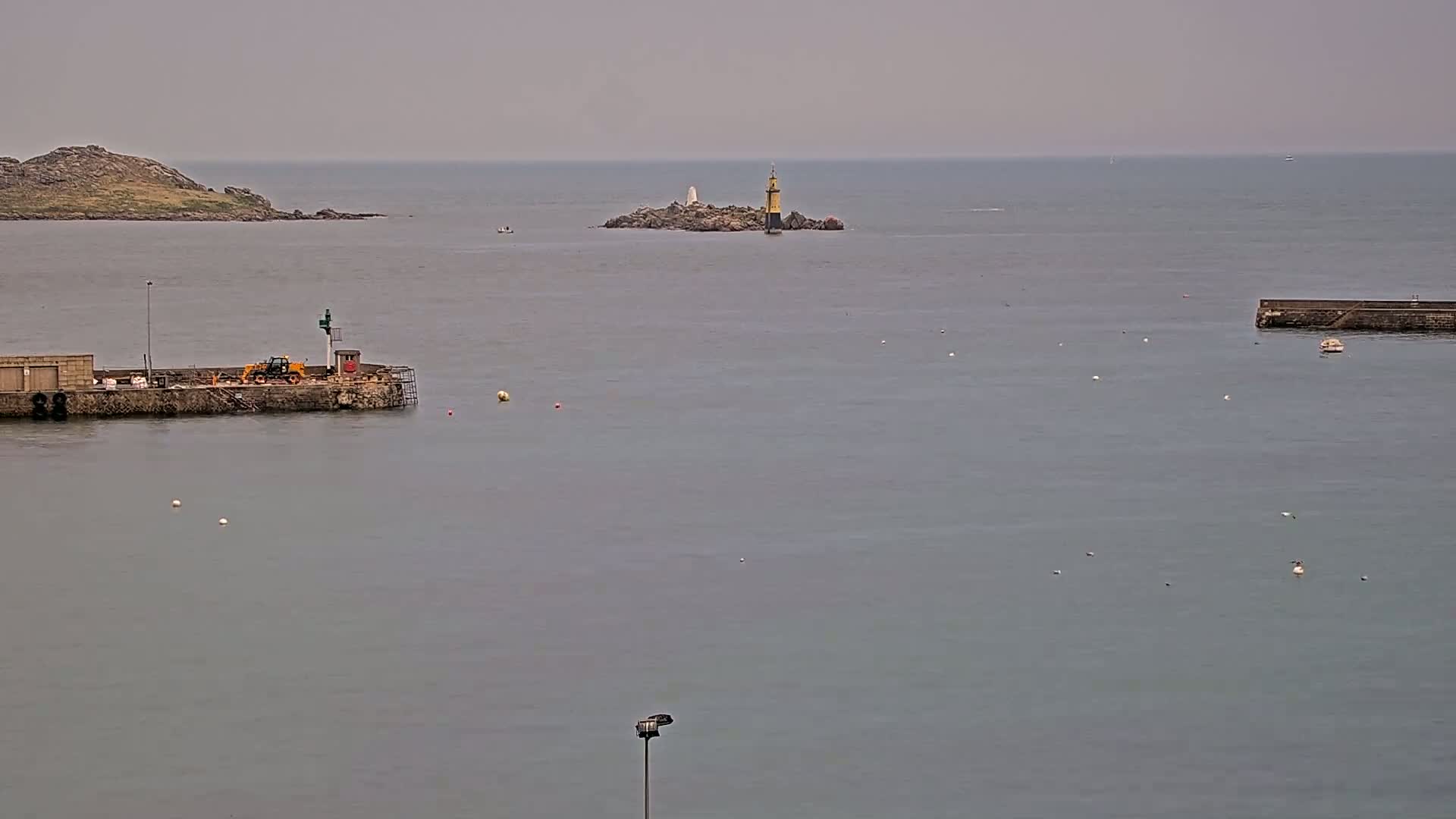 A small island with a lighthouse is visible in a calm, gray sea, near a harbor with a small piece of land that has construction equipment.