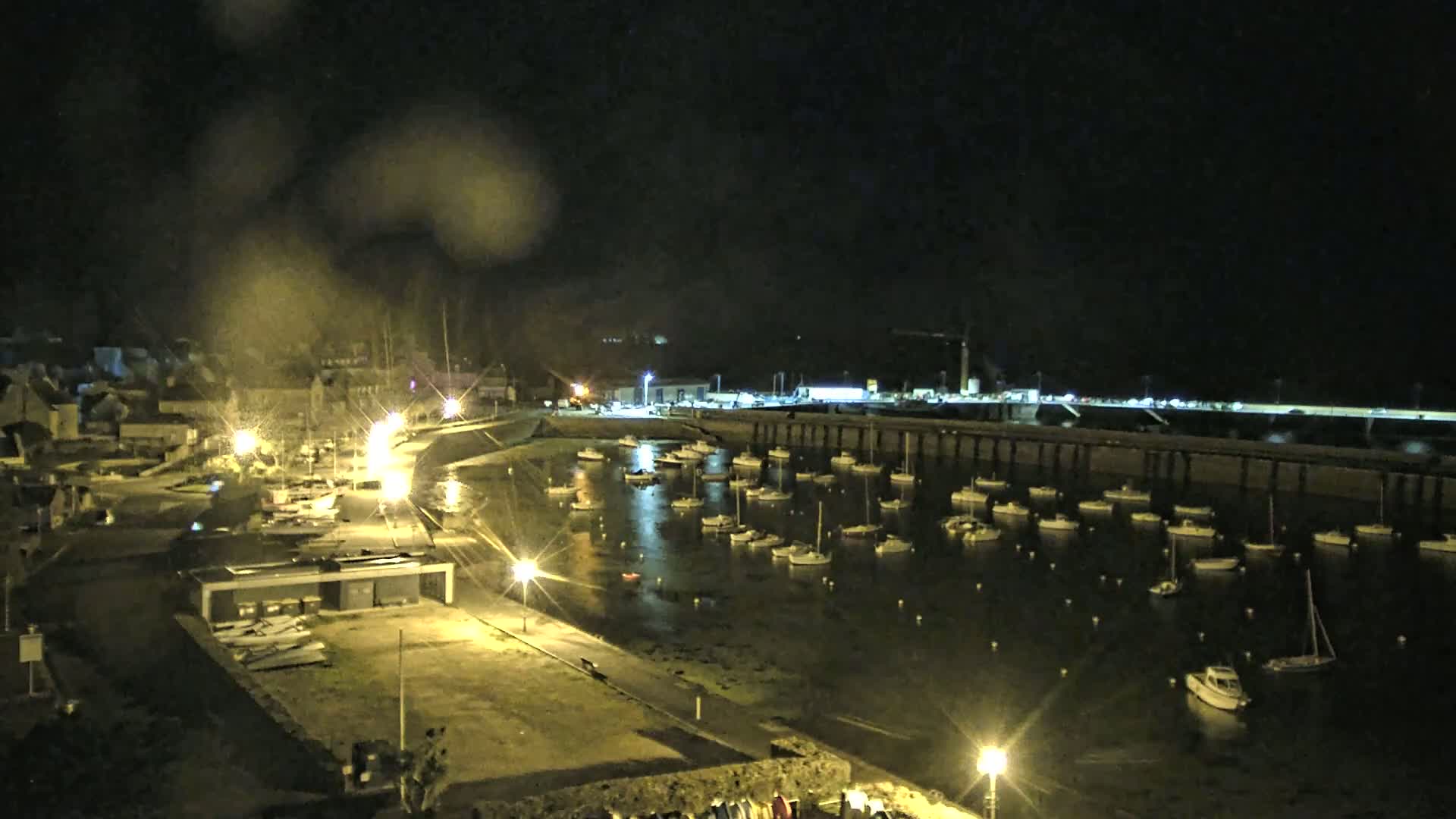 A nighttime view of a harbor filled with boats, seen through rain-covered glass, is lit by streetlights along a paved waterfront.