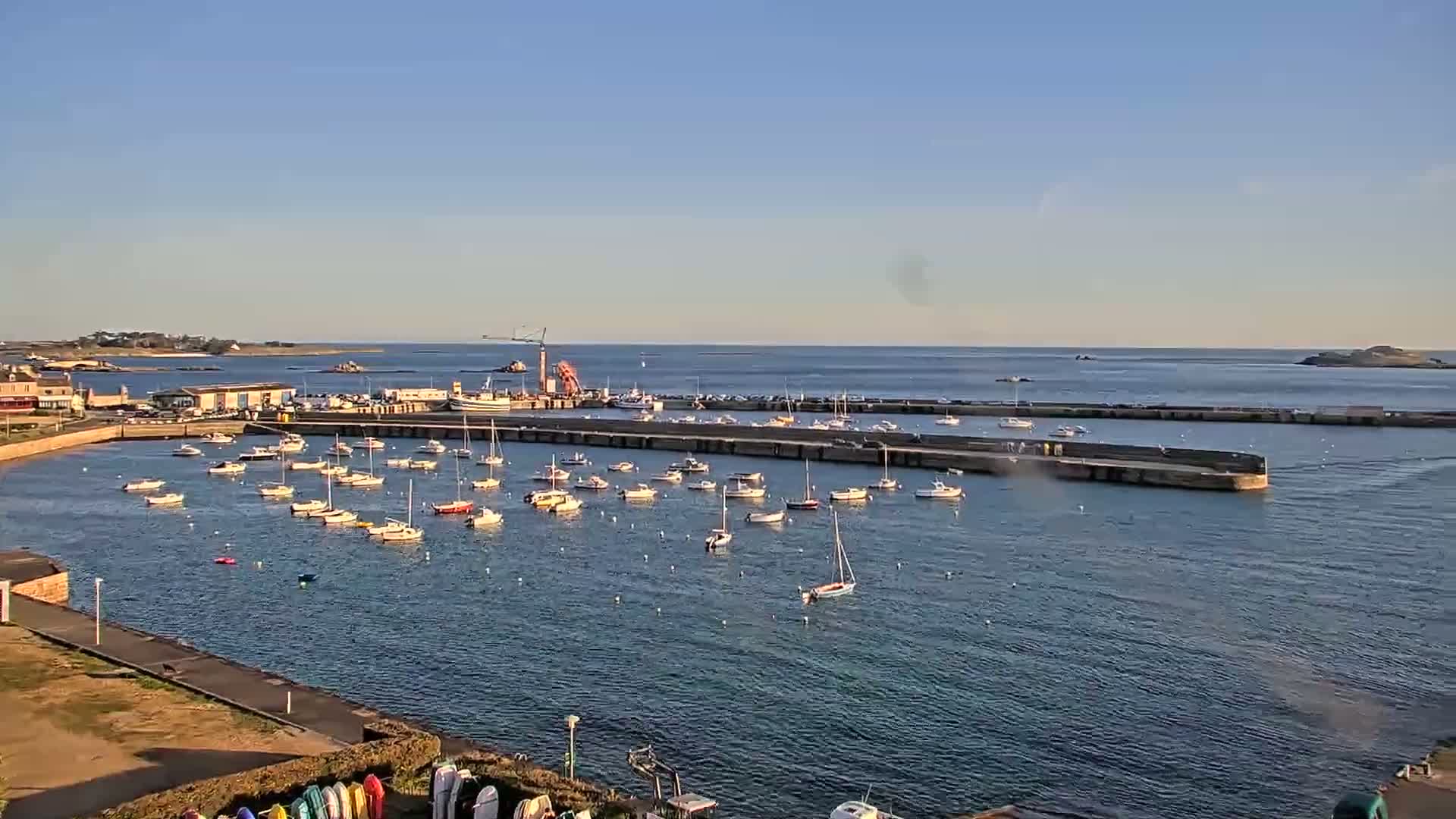 A harbor filled with numerous sailboats and small motorboats is seen under a clear, sunny sky.