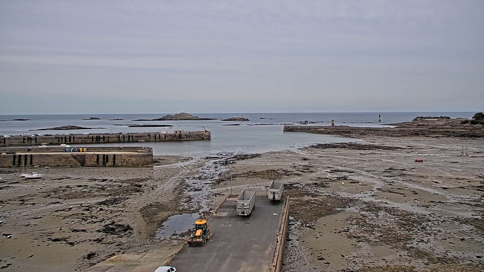 A harbor with a low tide exposing a wide expanse of mudflats, two trailers, and a small bulldozer are near a stone harbor wall, under an overcast sky.