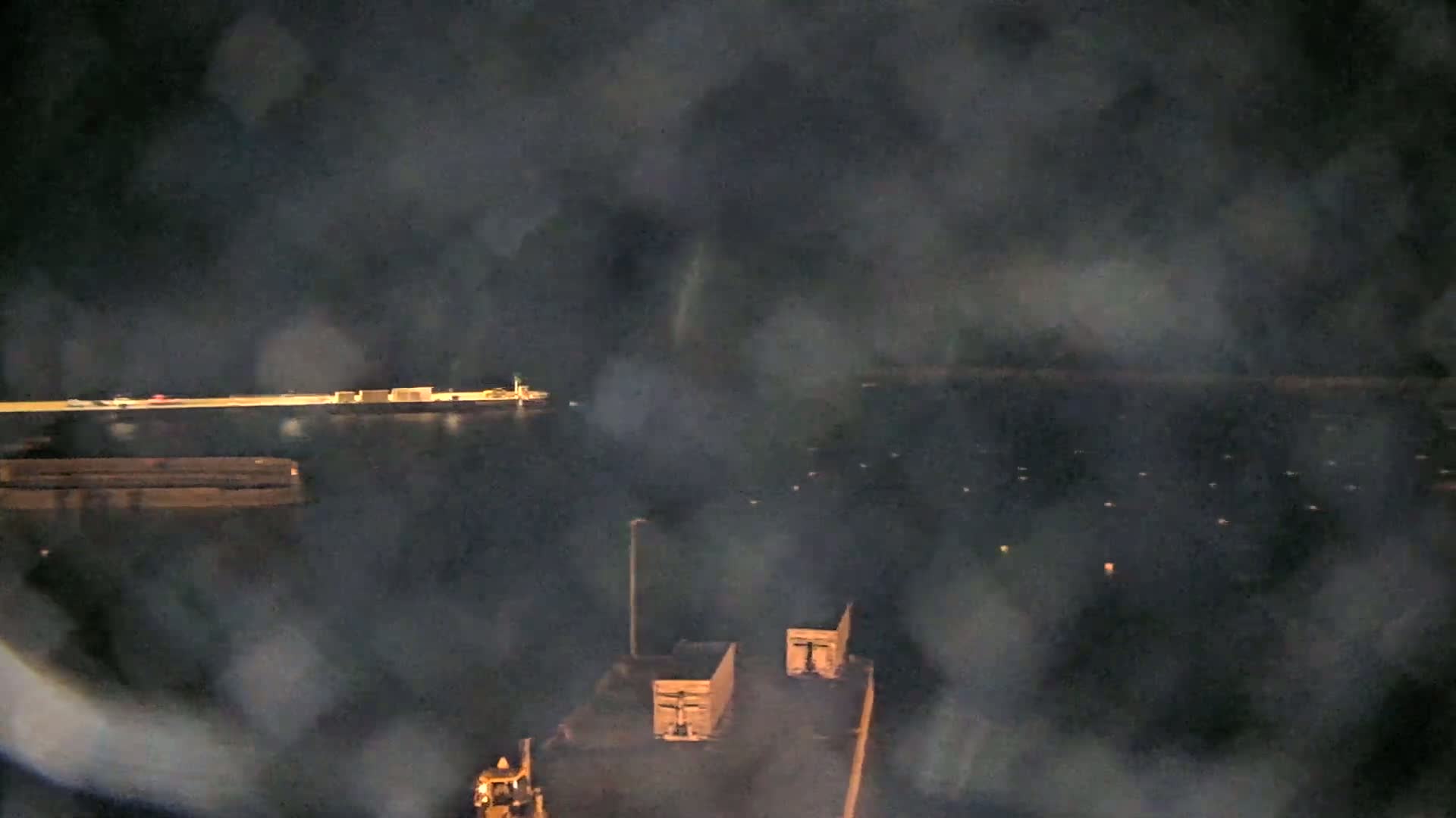A nighttime view of a harbor shows a body of water with a ship in the distance, some structures on a dock, and several pieces of industrial equipment in the foreground under a hazy or foggy sky.