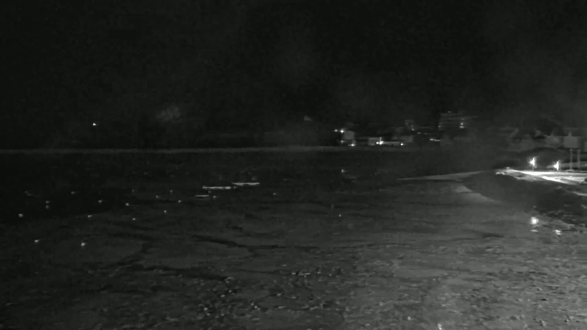 A dark, nighttime view shows a body of water reflecting a few distant lights, with a shoreline and dimly lit buildings visible in the background.