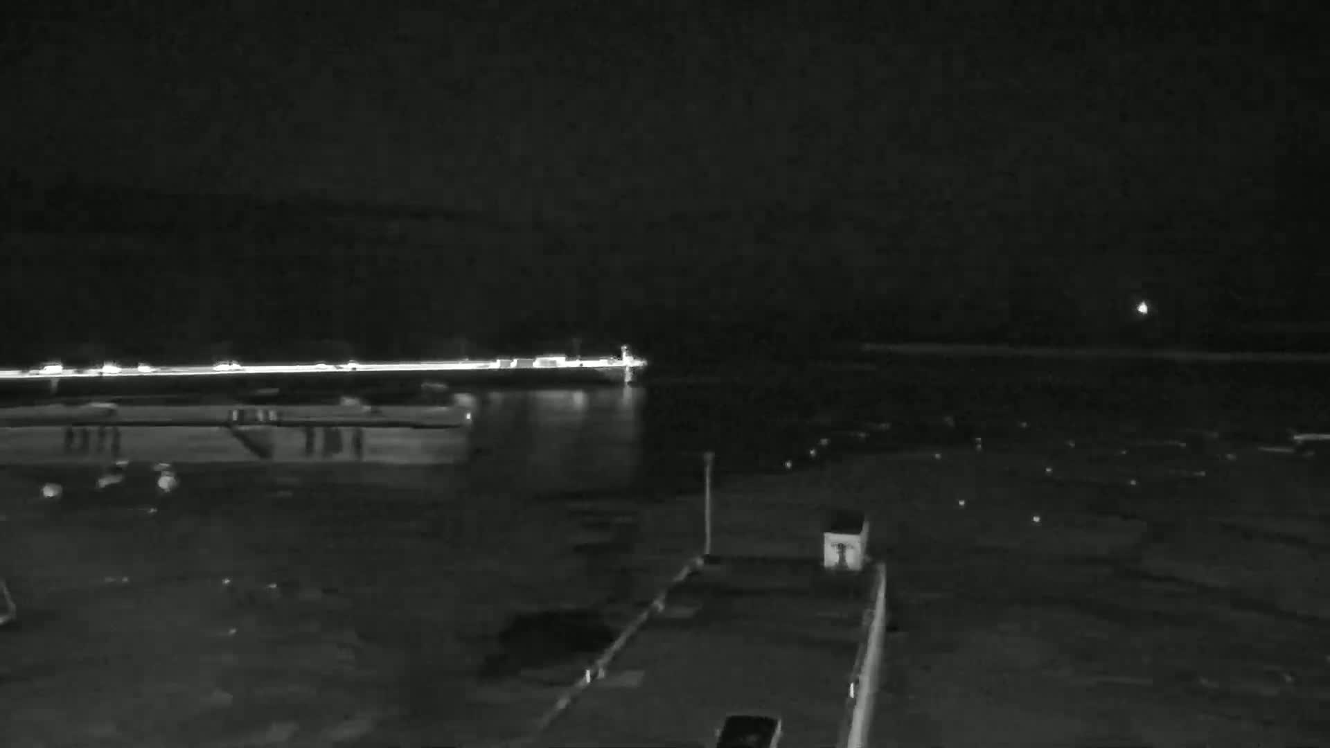 A nighttime view of a calm body of water with a line of lights in the distance, and a dark, empty dock in the foreground.